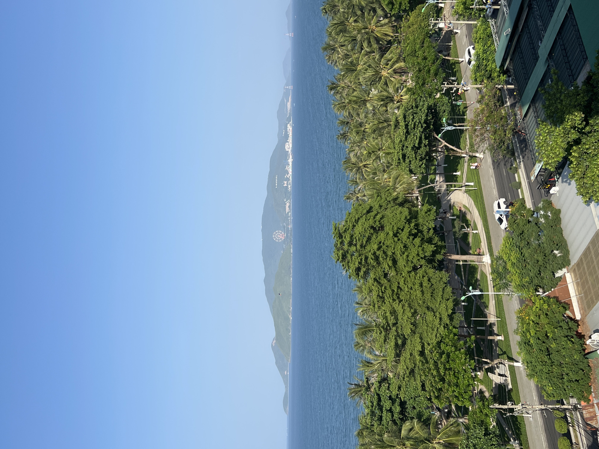 This is an eye-level, long-shot, vertical, outdoor photograph taken from a high vantage point, looking towards the east over a coastal city in Nha Trang, Vietnam. The scene depicts a clear, bright day with a vibrant blue sky dominating the upper portion of the frame.
In the mid-ground, a wide expanse of calm, deep blue ocean stretches towards a distant, green, hilly island chain. The coastline of the island is dotted with buildings and what appears to be a Ferris wheel, suggesting a developed tourist area. A few boats are visible on the water, and one appears to be a small lighthouse structure.
In the foreground and mid-ground, a dense line of palm trees forms a lush green border along the coast. Beyond the palm trees, a city street runs parallel to the ocean. The street is lined with trees, sidewalks, and streetlights. Several white cars are visible on the street, with one turning. A few people are walking along the sidewalk. The architecture of buildings is visible in the lower right corner, with a sloped, green roof.
The overall impression is one of a tropical coastal city with abundant greenery and a clear, sunny day. The image captures a scenic view of the ocean, coastline, and urban environment of Nha Trang.