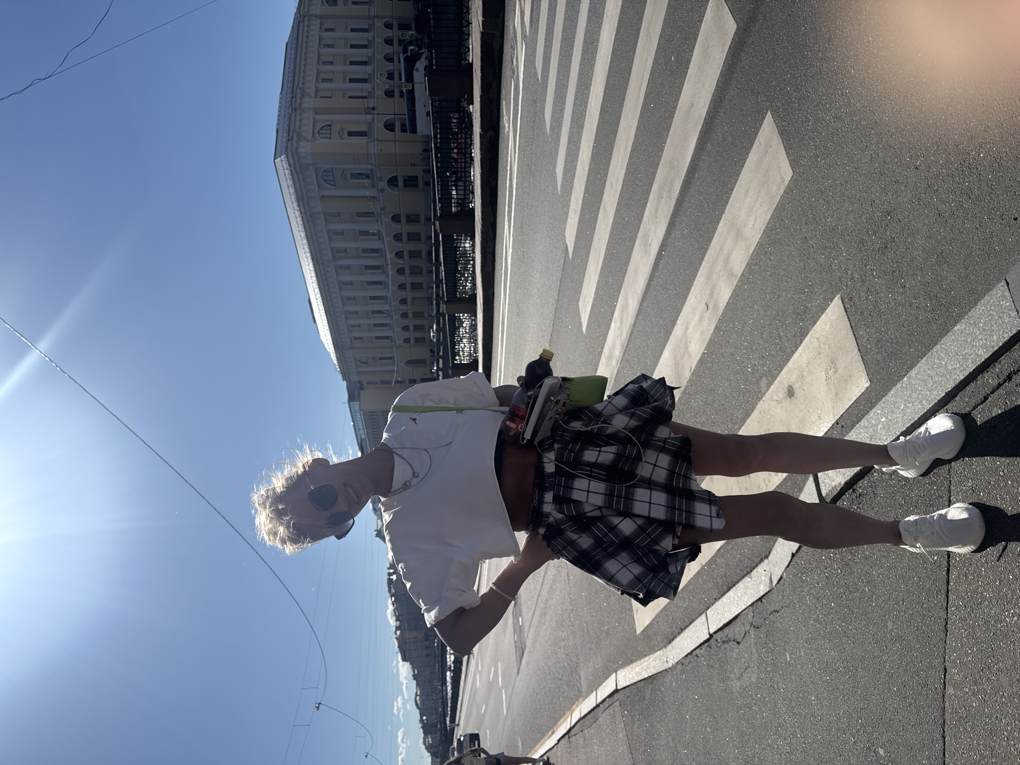 A person stands on a crosswalk in Saint Petersburg, Russia, under a clear blue sky. The sun is shining brightly, casting long shadows from the person and the white stripes of the crosswalk. The person is wearing sunglasses, a white crop top, and a black and white plaid skirt. They are also wearing white sneakers. A green bag is slung over their shoulder. In the background, a large, ornate building with multiple windows and columns is visible. Wires are strung across the sky, and a few cars are visible in the distance. The overall scene suggests a sunny day in a city, with the person enjoying the outdoors. The time of day appears to be midday, given the strong sunlight and shadows. The weather is clearly fair. There is no visible text in the image.