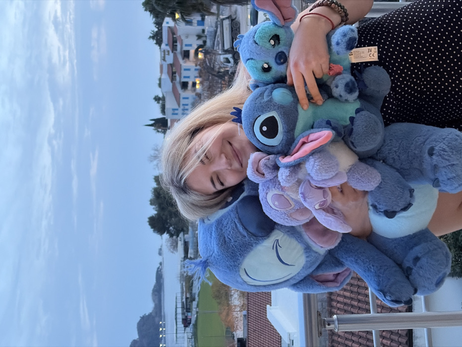 A blonde-haired female, smiling with eyes partially closed, is captured from the chest up, embracing five *Lilo & Stitch* character plush toys. A large light blue Stitch plush is positioned on her left, while various smaller blue and purple Stitch and Angel plush toys are held towards her right. A product tag is visible on the smallest blue Stitch plush. She is wearing a dark, white-polka-dotted garment and a beaded bracelet on her right wrist. The scene is situated on an elevated outdoor area, likely a balcony or terrace, in Budva, Montenegro. The background on the left reveals a bay or sea with a curved coastline, illuminated coastal structures, a green field, and a distant landmass. To the right, characteristic Mediterranean-style white buildings with blue window shutters and red-tiled roofs are visible amidst trees, including cypress and palm varieties. A construction dump truck is partially discernible on a road below the buildings. The sky is overcast and light blue.