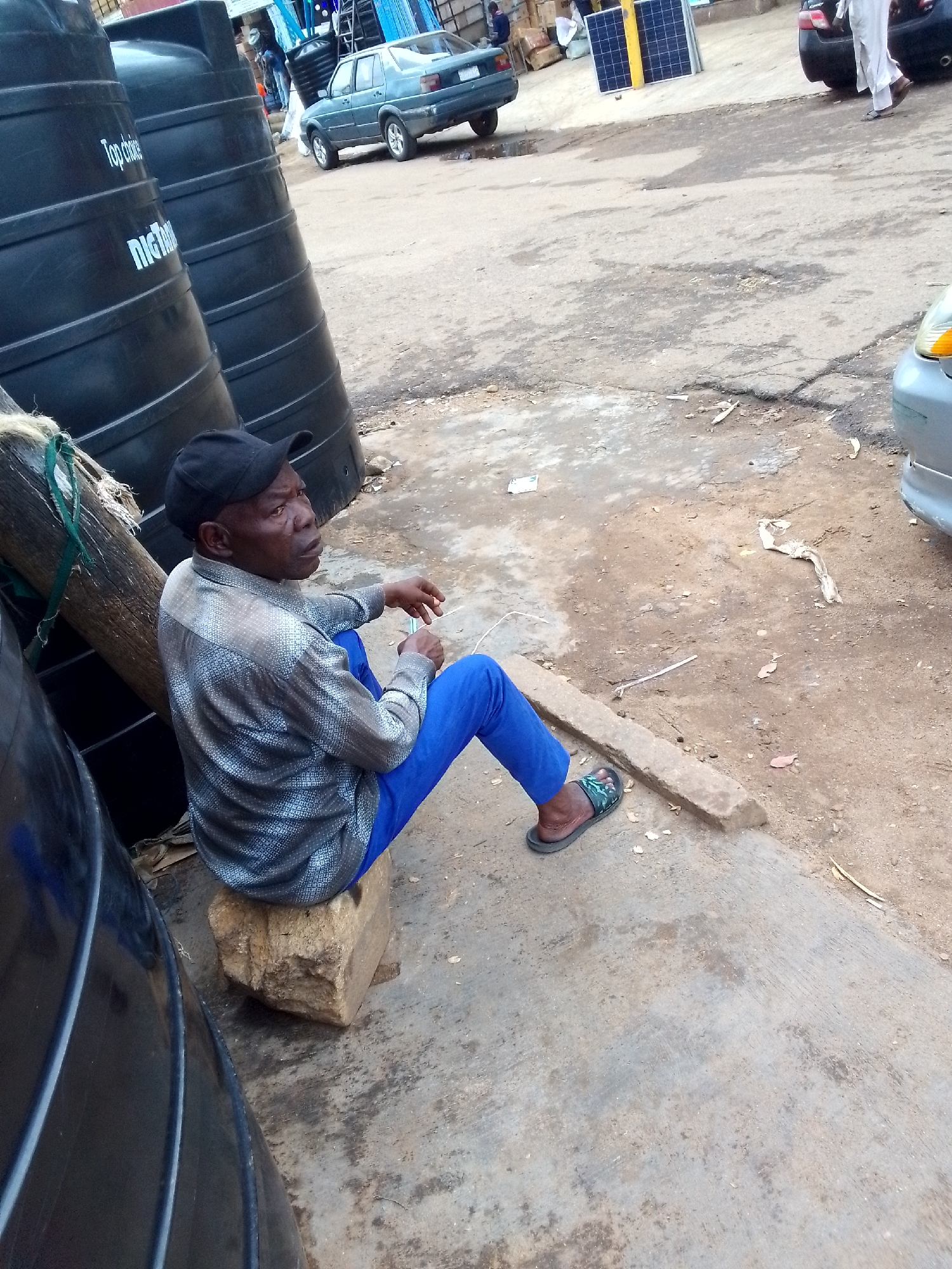 An African man wearing a black baseball cap, a patterned button-down shirt, and blue pants sits on a light-colored rock on a dusty street. He is looking to his right with his hands resting on his lap. Behind him, to his left, are several large black water tanks, one with "Top choice" and "NiCTANK" printed on it. To the right of the man, a teal sedan is parked on the street. In the background, a person in white clothing walks away from the camera. Further in the background, there are solar panels and boxes stacked. The ground is a mix of cracked concrete and dirt.