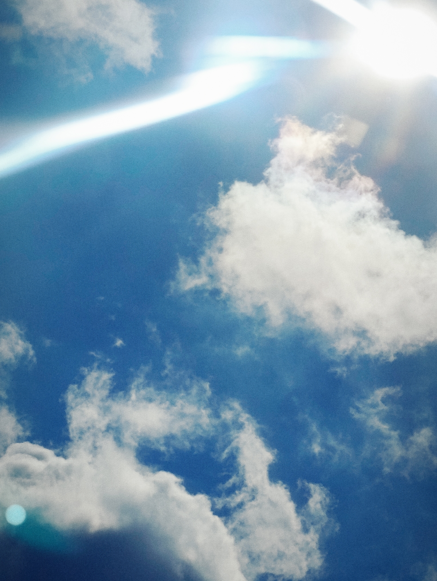 The image captures a bright sky with scattered white clouds and a strong sun glare. The sun appears to be partially obscured by a cloud or atmospheric haze, creating a prominent lens flare that streaks across the upper left portion of the frame. The sky is a vibrant blue, and the clouds vary in density, with some appearing wispy and others more solid. There is a noticeable blue circular artifact in the lower left quadrant, likely a reflection or lens anomaly. The overall scene suggests an outdoor, daytime environment with clear weather conditions, possibly a bright, sunny day. There are no visible people or man-made structures.