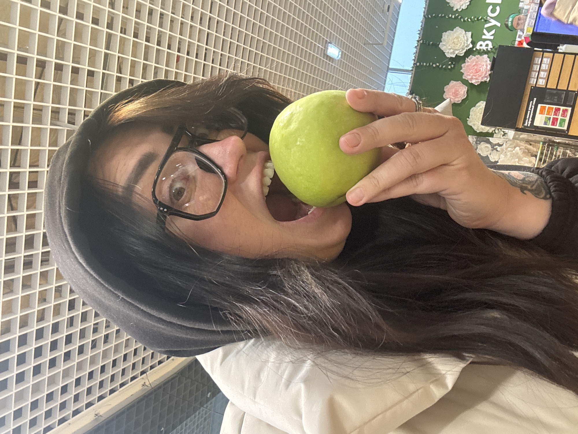 A woman with dark hair and black glasses is holding a green apple up to her mouth, as if she is about to bite it. She is wearing a black hoodie and a white jacket. The background consists of a white grid pattern and a floral display with the word "Вкусно" visible. The location appears to be a store, likely in a city, but the specific city is unknown.