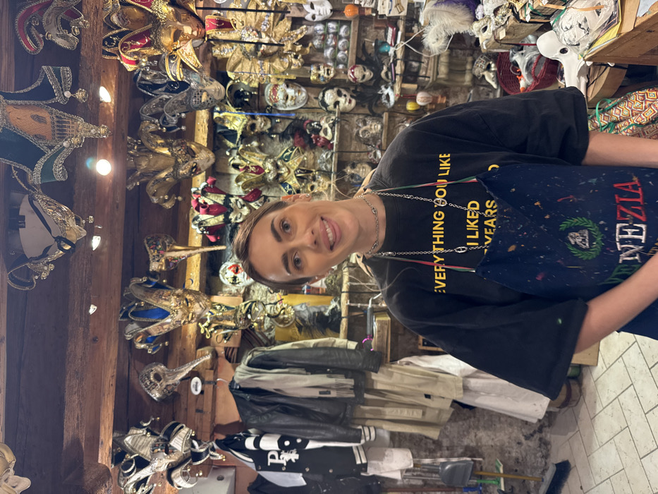 A single adult female is depicted from the waist up, smiling directly at the camera, inside a retail establishment in Venice, Italy. She has light brown hair pulled back and wears a black long-sleeve shirt with yellow text reading "EVERYTHING YOU LIKE" and "I LIKED YEARS" (partially visible), along with a paint-splattered dark blue apron embroidered with a gondola emblem and "VENEZIA ITALIA". Two silver-colored necklaces are visible on her neck. The background is densely filled with hundreds of elaborate, traditional Venetian carnival masks displayed on walls and hanging from overhead wooden beams. Mask types include Bauta, Columbina, Volto, Jester, and Medico della Peste designs, adorned with feathers, gold accents, and various colors. To the left, several garments, including a black leather jacket and a beige trench coat, hang on a rack. The environment features exposed wooden beams and tiled flooring, consistent with an artisan workshop or mask shop.