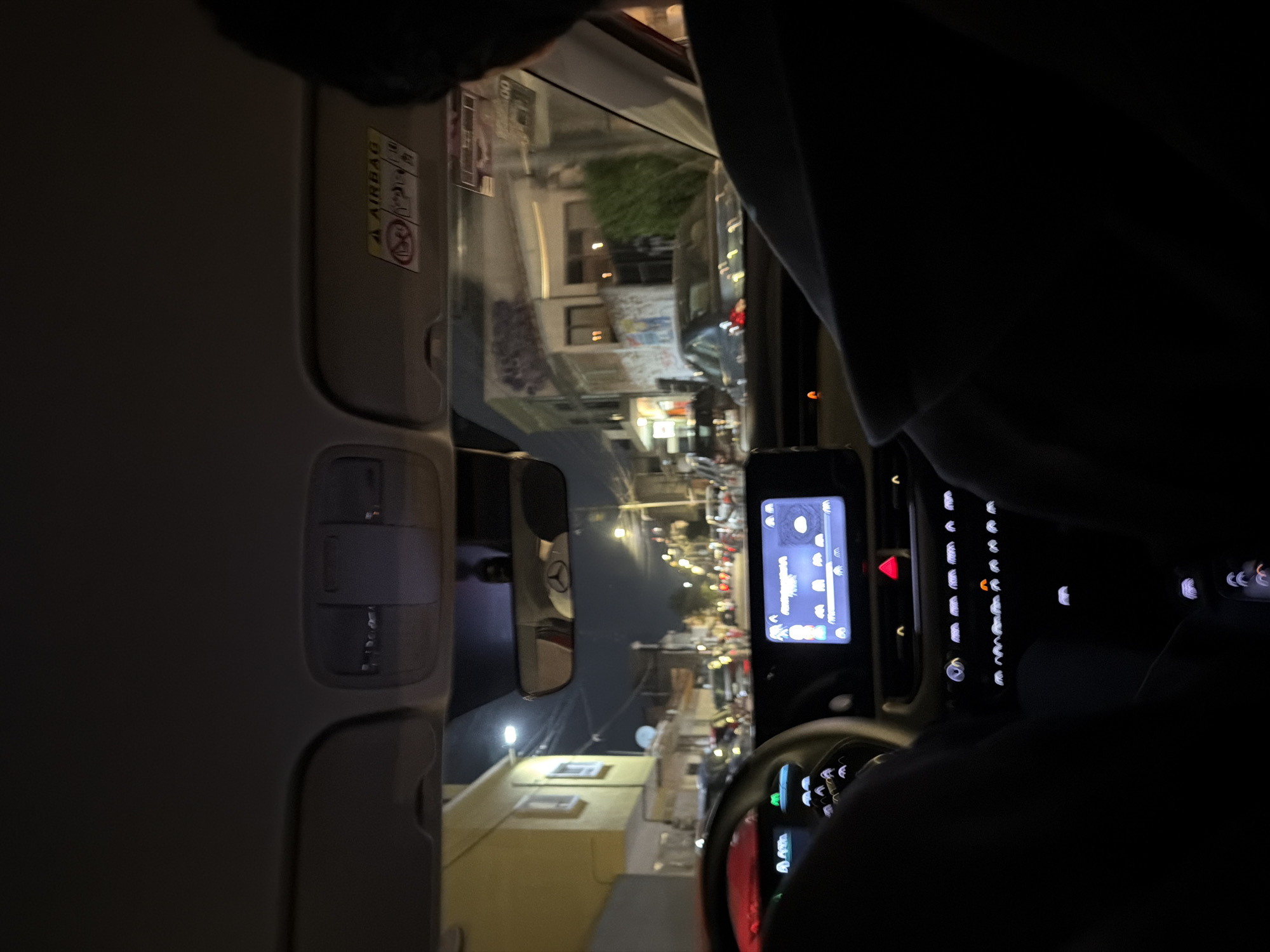 The view from inside a car in San Andrés Cholula, Mexico at night. The interior of the car is visible: the steering wheel, center console with a screen displaying information, and the sun visor. Outside the window, street lights illuminate buildings and traffic on the street. The buildings appear to be low-rise, with some having illuminated windows. There is traffic including cars and possibly motorcycles moving down the street.