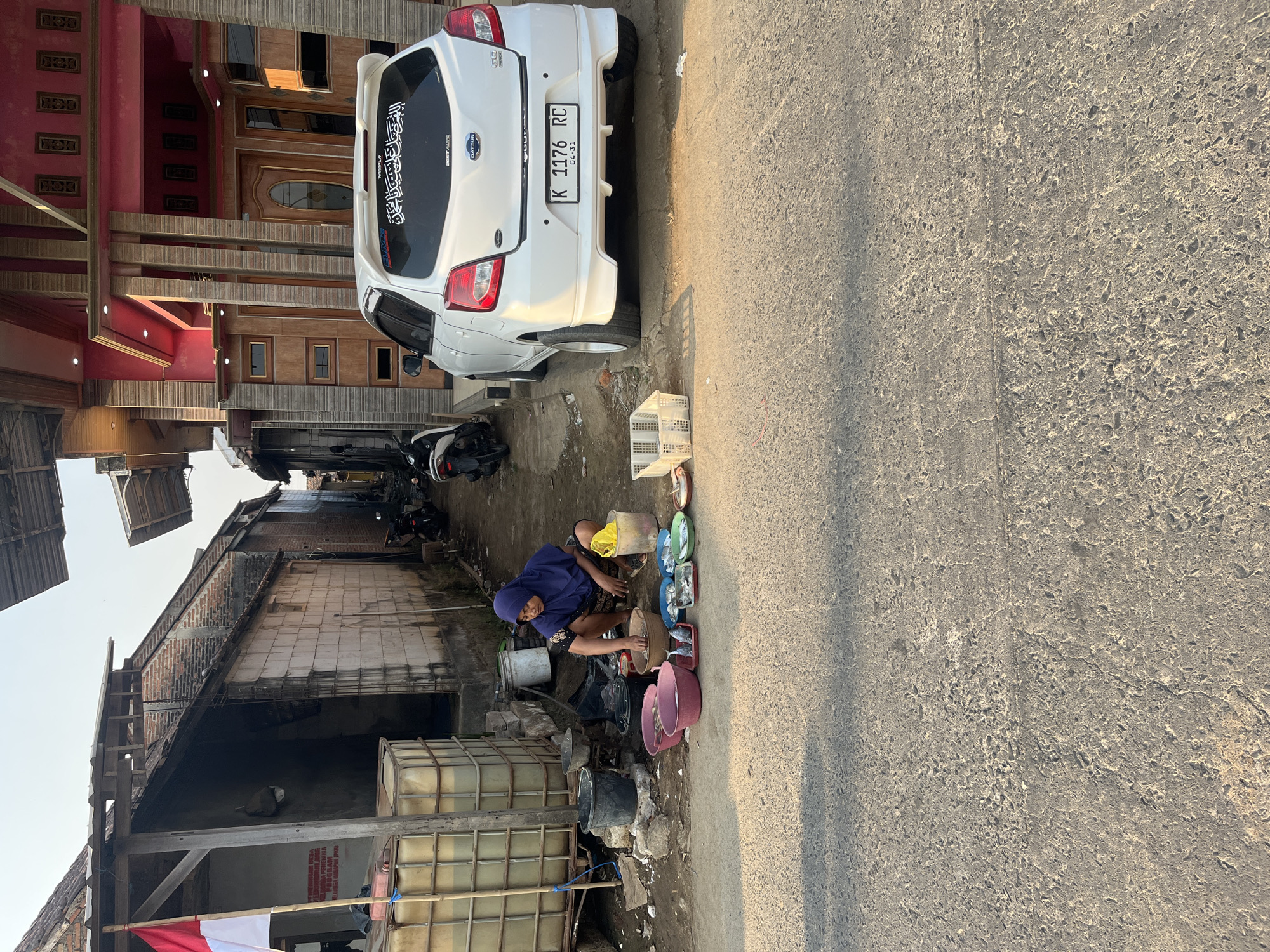 A woman wearing a blue hijab and patterned skirt is crouching on the ground in front of a row of food items arranged on the pavement. The items are in various bowls and plates, appearing to be fish. She is holding something in her right hand, possibly preparing the food. Behind her, to the left, is a large plastic tank and a small building with a red and white flag. To her right, a white car is parked on the street, and further to the right, a modern house with a red facade and wooden accents is visible. The scene appears to be a street in a residential area, possibly a market stall set up outdoors. The lighting suggests it is either early morning or late afternoon. The overall environment is somewhat cluttered with various objects and buildings indicating a mixed urban and rural setting.