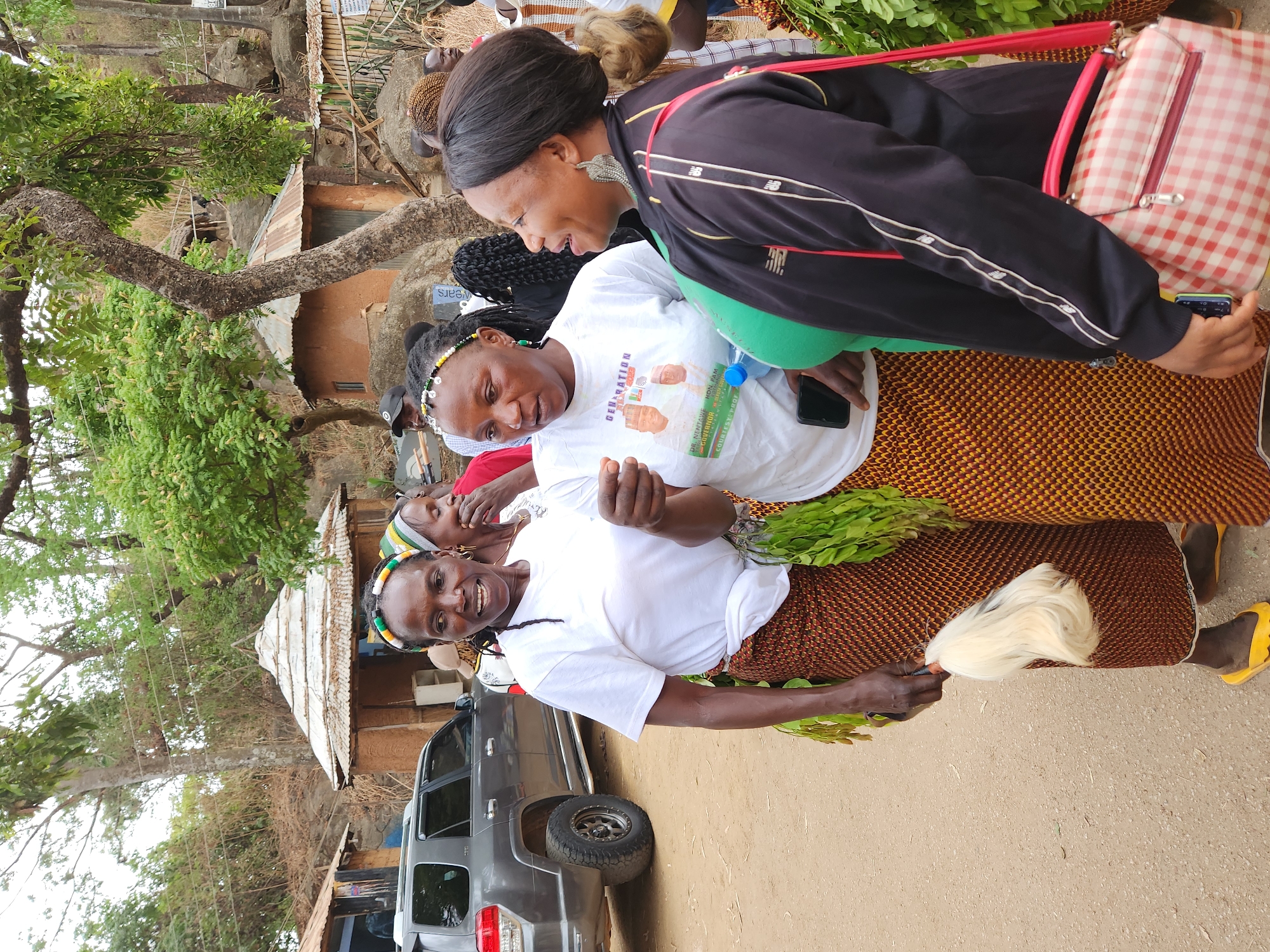 The image depicts an outdoor scene in Jos, Nigeria, during daylight hours, showing several women gathered in what appears to be a community setting.
In the foreground, a woman wearing a white t-shirt, a colorful patterned skirt, and a beaded headband smiles towards the viewer. She holds a bundle of green leafy branches in one hand and a white, fluffy, traditional-looking object, possibly a fly-whisk, in the other. Adjacent to her, another woman wears a white t-shirt with a political campaign message: "GENERATION" and "DR. MERTARUE, HON. PAM GOVERNOR," accompanied by images of two men. She holds a blue water bottle and a smartphone. A third woman, wearing a black jacket over a green top and carrying a red and white plaid shoulder bag, is seen interacting with the others, smiling.
The background features a grey SUV parked on unpaved ground. Simple structures with corrugated iron roofs are visible among sparse vegetation, trees with prominent trunks, and rocky terrain, suggesting a rural or semi-urban environment. Another partial word, "WEARS," is visible on a sign or poster in the background. The overall atmosphere suggests a communal gathering or event, possibly related to a political campaign given the text on one of the t-shirts.