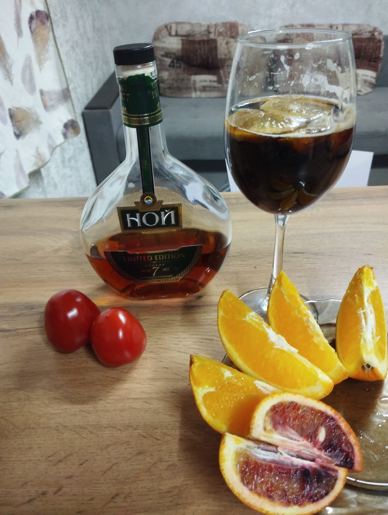 The image captures an indoor still life arrangement featuring a bottle of brandy, a drink in a glass, and various fruits on a wooden table.
The central subject is a clear glass bottle of "НОЙ" (Noy) brandy, which is a "LIMITED EDITION" and aged "7 ЛЕТ" (7 years), identified as "КОНЬЯК АРМЕНИИ" (Cognac of Armenia) on its label. The bottle contains a amber-colored liquid. To its right, a stemmed wine glass holds a dark beverage, possibly a cocktail, with several thin slices of what appear to be dried or fresh lemon or orange floating on top. Some froth or residue is visible on the inside of the glass rim.
Arranged on the wooden surface are several fruits. Two bright red, oval-shaped cherry tomatoes are placed near the base of the brandy bottle. Further to the right, on a small, round, silver-colored coaster or dish, are several slices of orange. One orange has been cut in half horizontally, revealing its distinct dark red, almost purplish flesh and seeds, indicating it is a blood orange. Regular orange wedges are also present.
The background shows a soft-focus interior, with a light-colored curtain or fabric draped to the left, featuring abstract patterns resembling feathers. A piece of furniture, possibly a sofa or chair with patterned upholstery, is partially visible in the upper right background. The setting appears to be a cozy indoor environment, likely a home in Grishanki, Russia, as suggested by the provided context. The lighting is soft and ambient, without any strong shadows indicating a particular time of day. No people are visible.