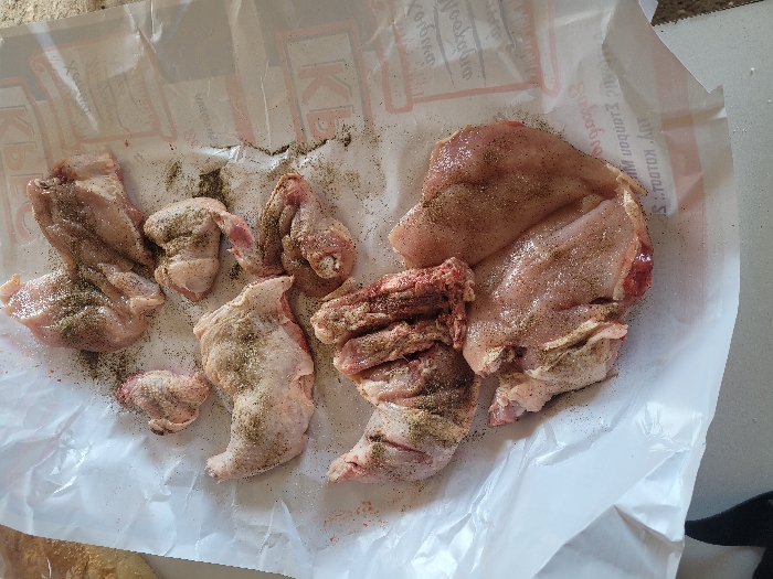 The scene captures a close-up, overhead view of raw, butchered chicken pieces seasoned with black pepper and salt, arranged on a sheet of white butcher paper. The paper prominently displays red and grey Greek text, including "ΚΡΕΟΠΩΛΕΙΟ" and "ΑΝΔΡΑΒΙΔΑ", identifying the location as Ανδραβίδα, Greece. Visible chicken components include bone-in and boneless portions, such as a thigh, breast cuts, and wings, exhibiting pink flesh and some skin. Dark specks of ground pepper and fine white granules of salt are distributed across the chicken surfaces and the surrounding paper. A blurred, light-brown object, possibly bread, is partially visible in the upper-left corner. The butcher paper rests on a light-colored, reflective surface, likely a kitchen countertop. No individuals or active interactions are present in the frame.