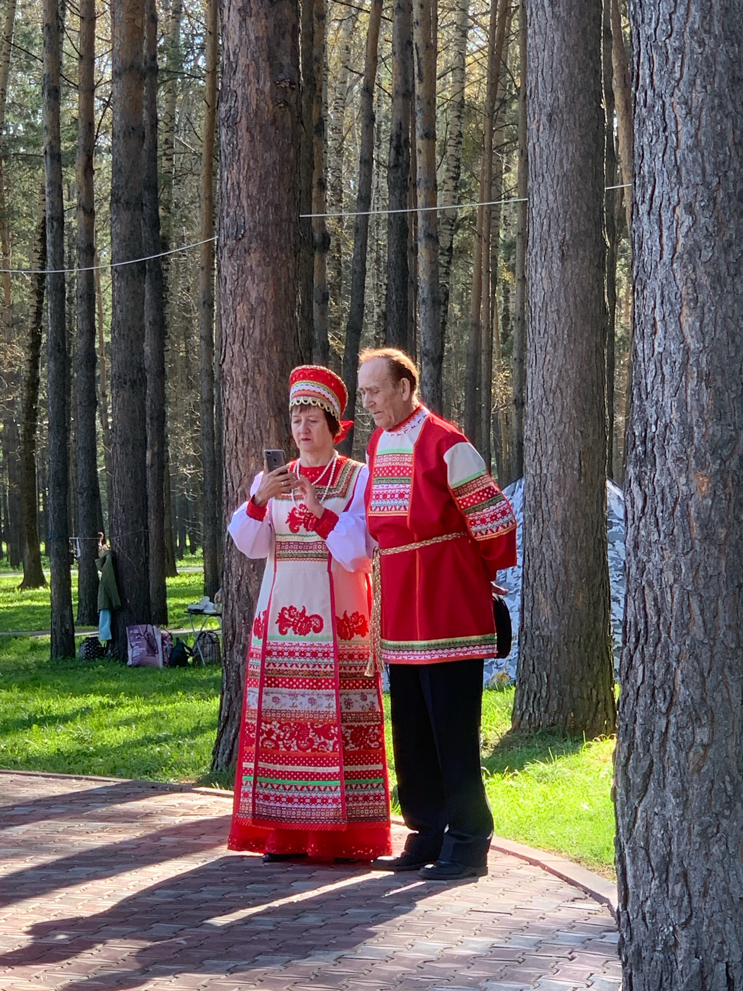 In Prokopyevsk, Russia, a man and a woman stand in a sun-dappled forest clearing. Both are dressed in traditional Russian attire. The woman, on the left, wears a red and white embroidered sarafan over a white blouse with red embroidery and a matching headdress. She is holding a smartphone and looking at its screen. The man, on the right, is in a red embroidered shirt with a braided belt and black trousers. He is looking to his left. Behind them, tall pine trees create a dense forest backdrop. The ground is a paved walkway with a patterned brick surface, casting long shadows. To the left of the couple, a grassy area with some bags and a person in a green jacket can be seen.