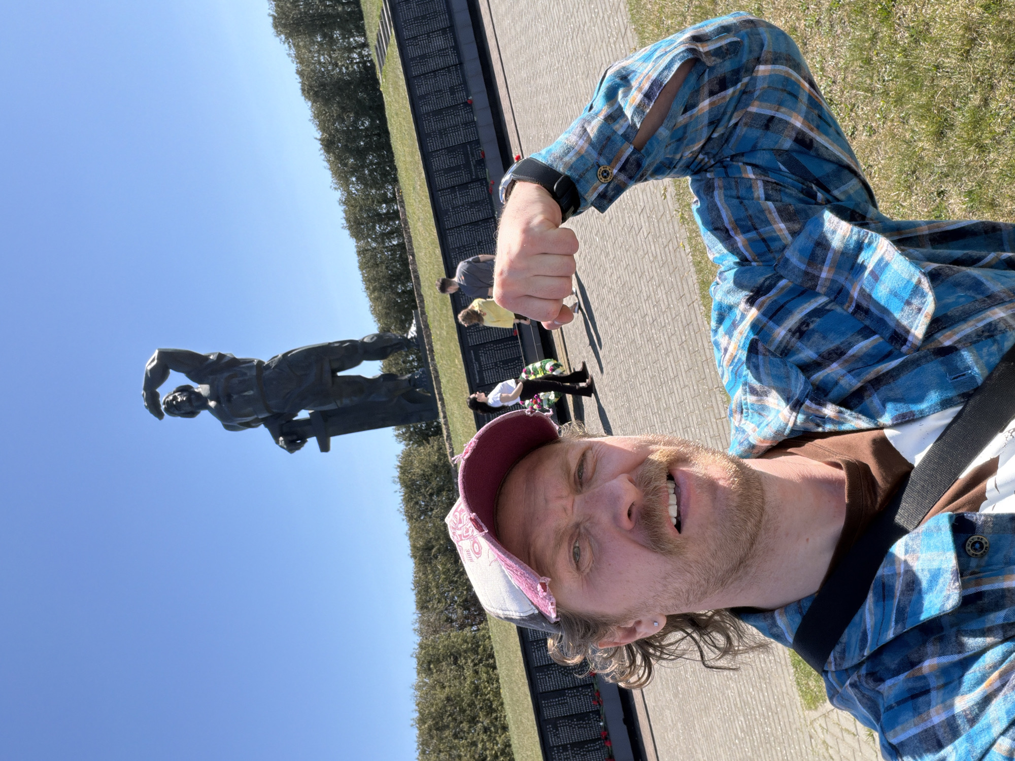 A man is taking a selfie in front of a large monument, likely a war memorial, in Свіслацкі сельскі Савет, Belarus. The man is wearing a blue plaid shirt with cut-out sleeves and a baseball cap, and is clenching his fist. The monument is a large bronze statue of a figure holding a sword or gun. Behind the statue is a wall with inscriptions, possibly names of those who died in the war, and a row of trees. Other people are visible in the background. The sky is blue and the weather appears to be sunny. The ground is paved.
