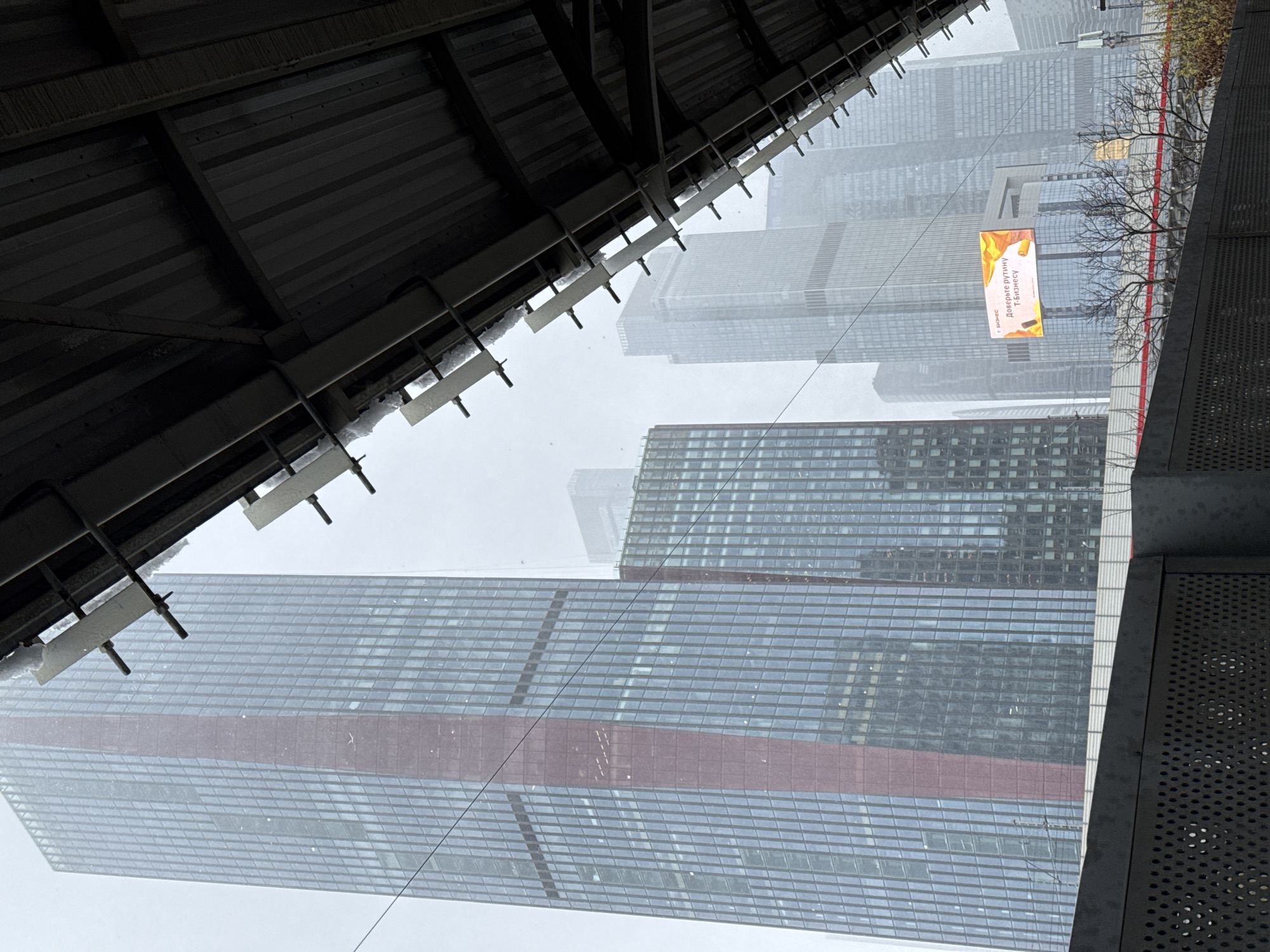 The image, taken during overcast weather conditions that might indicate snow or rain, shows a view of the Moscow International Business Center (Moscow-City) from an elevated position under a metallic structure, likely a covered walkway or elevated station. Several high-rise buildings are visible in the background, partially obscured by the weather conditions. The architectural styles of the buildings are modern, typical of a financial district. A horizontal advertising billboard is placed between buildings. A bare tree is near the billboard.