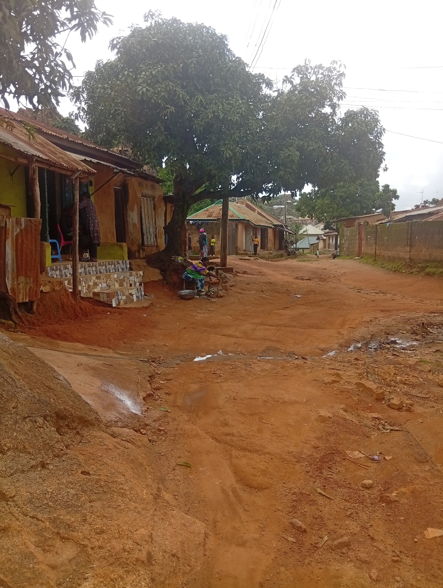 The image depicts a dirt road in a residential area of Jos, Nigeria. The road is unpaved and shows signs of wear, with puddles of water and uneven terrain. Several structures line the road, appearing to be houses made of mud brick and corrugated iron roofing. In the foreground, a person is seated on the ground next to a large metal bowl, possibly engaged in some domestic activity. A few other individuals are visible in the background, including one standing near the entrance of a building. A large tree with dense foliage stands prominently in the middle of the scene, providing shade. The overall environment suggests a semi-rural or developing urban setting.