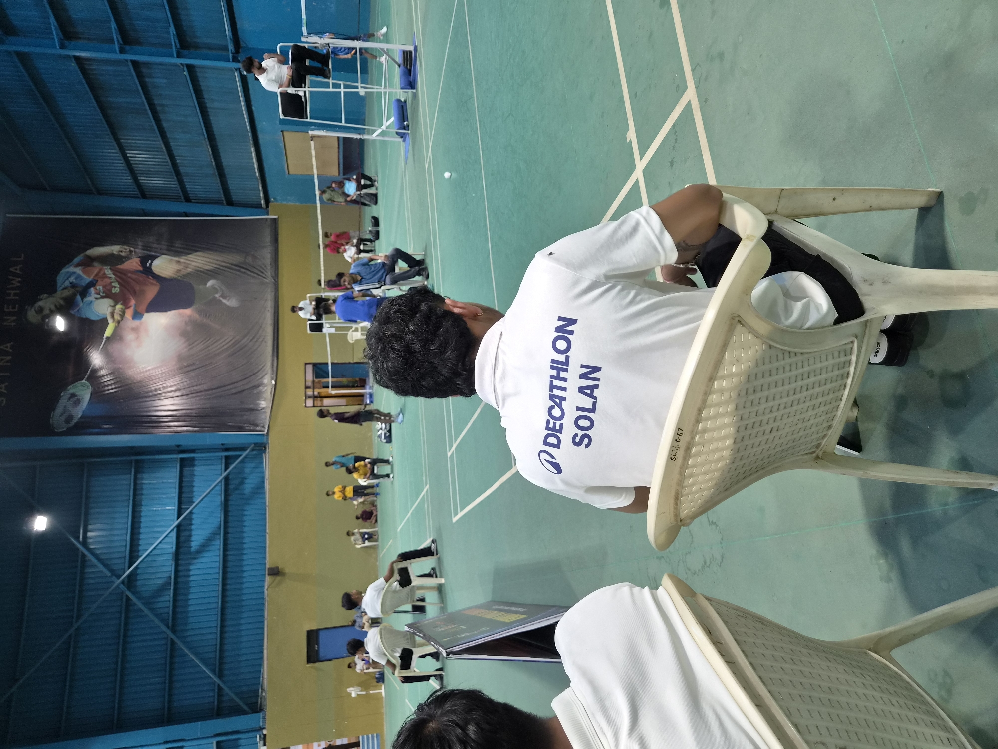 The image shows the interior of a badminton court in Phagwara, India. In the foreground, two individuals wearing white t-shirts are seated on plastic chairs, facing away from the camera, towards the badminton court. One of the t-shirts has "DECATHLON SOLAN" printed on the back. In the mid-ground, several people are present on and around the court. Two individuals appear to be playing badminton, with one in the action of hitting a shuttlecock. Another person is seated on a high umpire's chair. Spectators are also visible in the background, some seated and others standing. A large banner featuring a female badminton player is prominently displayed on the wall in the background. The court floor is green with white lines marking the boundaries. The overall environment suggests an indoor sports facility.