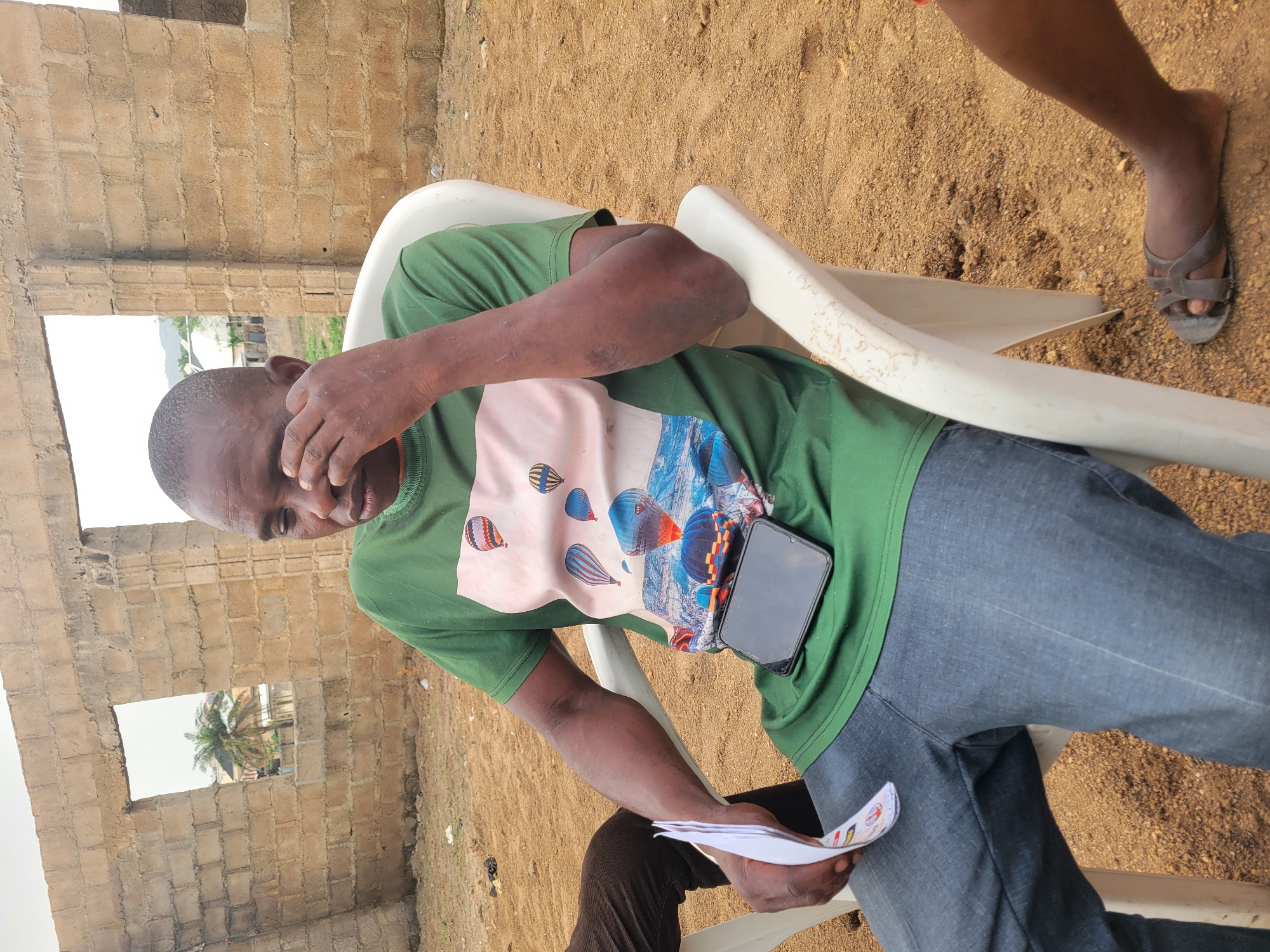 A man is sitting on a white plastic chair outdoors in Madalla, Nigeria. He is wearing a green t-shirt with a hot air balloon design, blue jeans, and brown sandals. He is holding papers in his left hand and covering his face with his right hand. A black smartphone rests on his lap. Behind him is a partially constructed brick building with open window frames. The ground is covered with brown dirt and sand. Another person's leg is partially visible on the right side of the image.