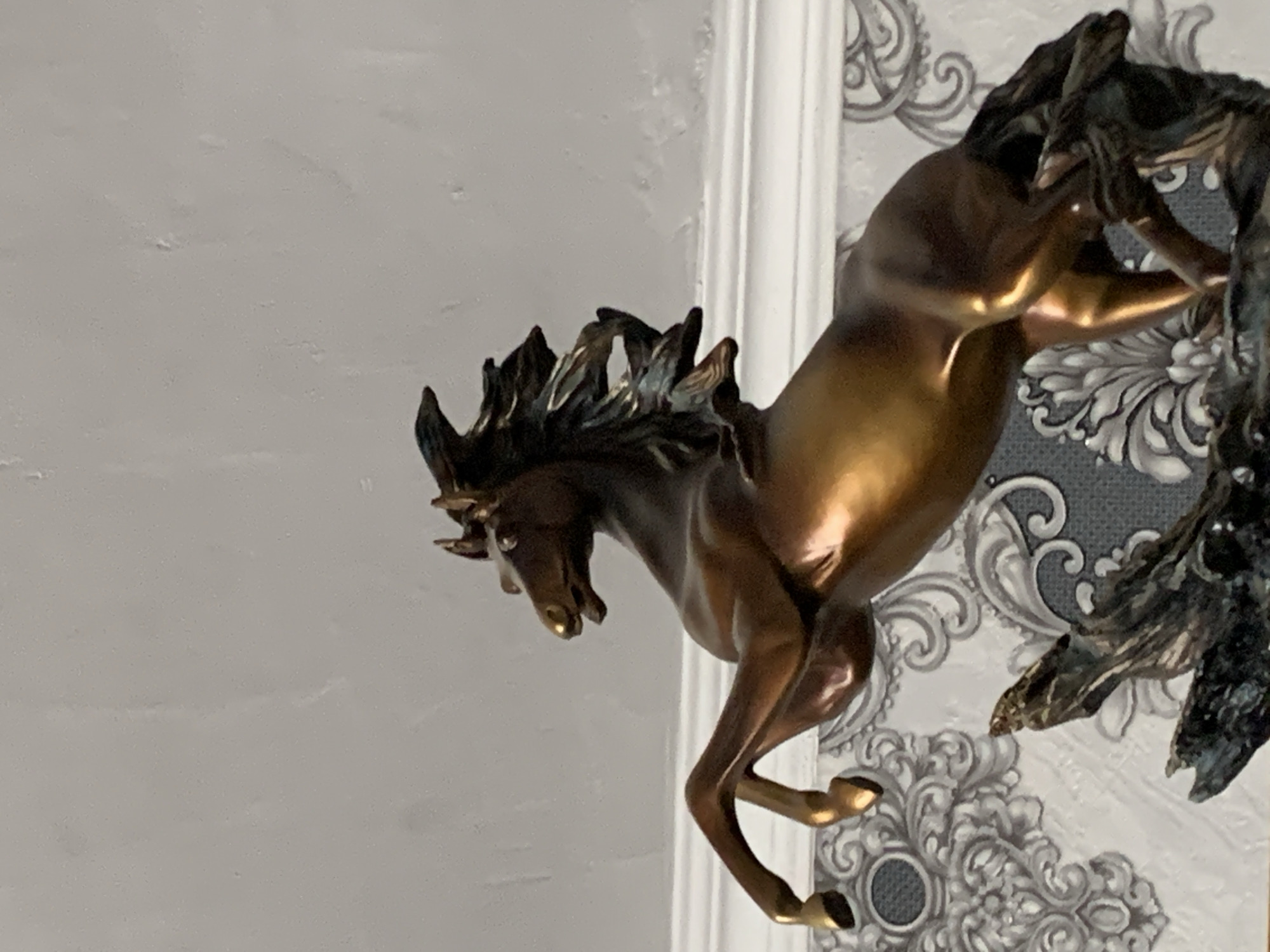 The image shows a bronze sculpture of a horse, posed as if rearing up on its hind legs. The horse is dark brown with golden highlights, and its mane and tail are flowing. It is positioned on a dark, rough-textured base that appears to be made of sculpted wood or rock. In the background, there is a wall with ornate wallpaper featuring a gray and white damask pattern. Above the wallpaper, there is white molding. The lighting suggests an indoor setting. The location of this sculpture within the city of Tashkent, Uzbekistan, cannot be determined from the image.
