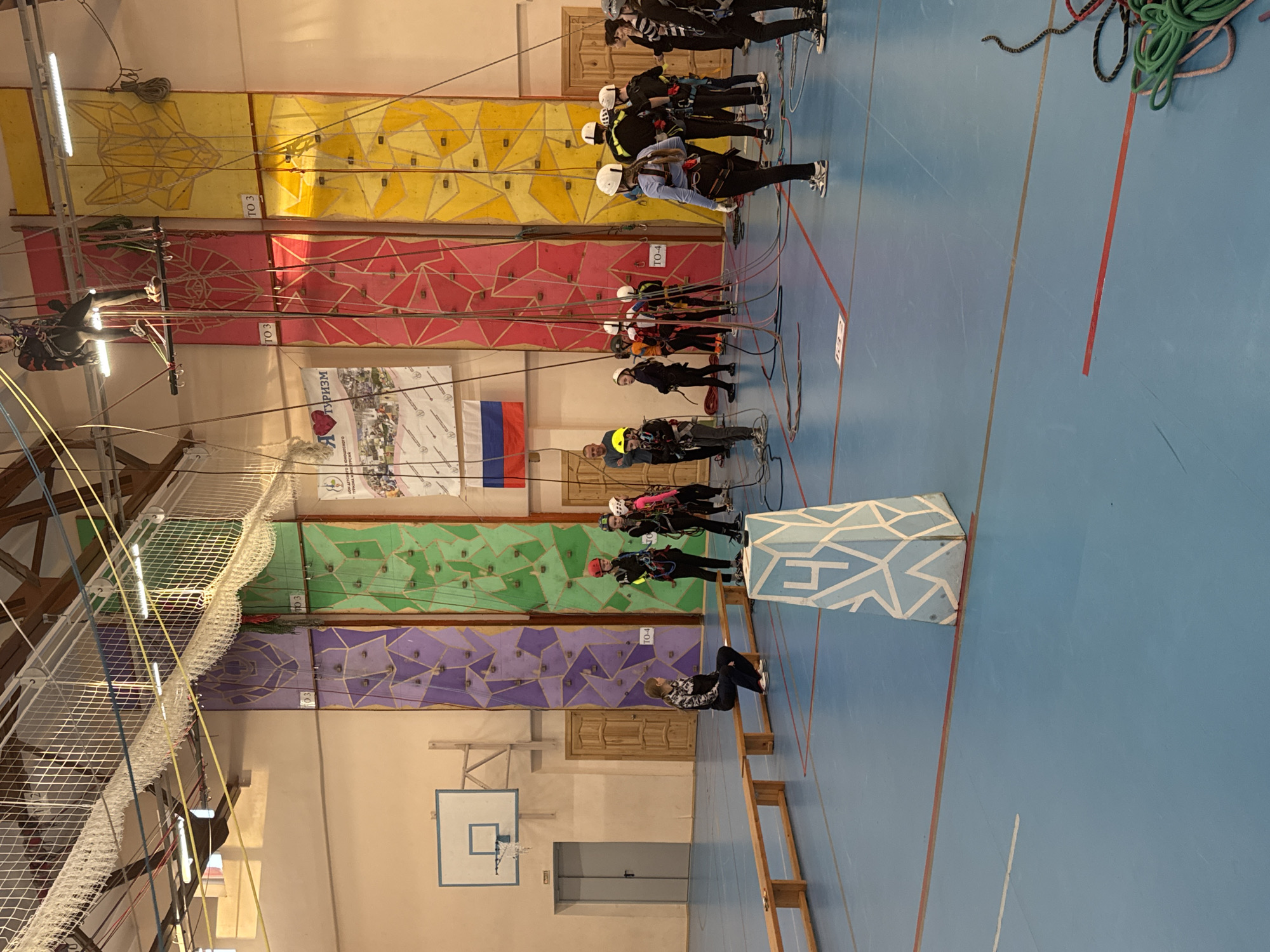 The image depicts an indoor climbing gym, likely in Berezniki, Russia, given the presence of the Russian flag. The gym features several climbing walls distinguished by color: yellow, red, green, and purple. People are standing at the foot of each wall, most wearing helmets and climbing harnesses and appearing to be connected to ropes. One individual is suspended from the top of the red climbing wall, presumably climbing.
The gym has a blue floor with red lines marking areas or paths. A wooden bench is present on the floor, near the purple and green climbing walls. A basketball hoop is mounted on the wall, and a white safety net is visible above the climbers. The scene suggests a training session or competition related to climbing sports.
