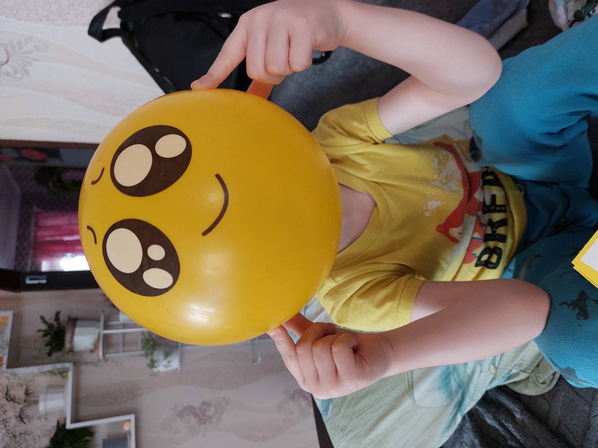 An indoor, close-up image shows a child lying down and playfully holding a large, bright yellow balloon that features a cheerful emoji face. The child's face is completely obscured by the balloon, which has distinctive black-outlined white eyes with black pupils, small black curved eyebrows, and a wide black smile. The child's fair-skinned hands gently grasp the top and bottom of the balloon.
The child is dressed in a short-sleeved yellow t-shirt, on which partial text is visible, including "RED" in red letters and "BK" in black, suggesting part of a word like "BRED" or "BREAK." They are also wearing light blue or teal sweatpants. The child's arms are bent, with one arm resting on a patterned light green and white fabric, and the other on a darker grey patterned fabric.
The background indicates a home environment. A dark backpack is visible behind the child's upper body, resting on a light-colored surface. Further back, a wall features light-toned, possibly floral or swirl-patterned wallpaper. White decorative shelves or frames, holding small green plants, are mounted on this wall. The lighting suggests daytime indoors. The image appears to be rotated 90 degrees clockwise. The setting is identified as Большой Букор, Russia. The scene conveys a lighthearted and playful mood.