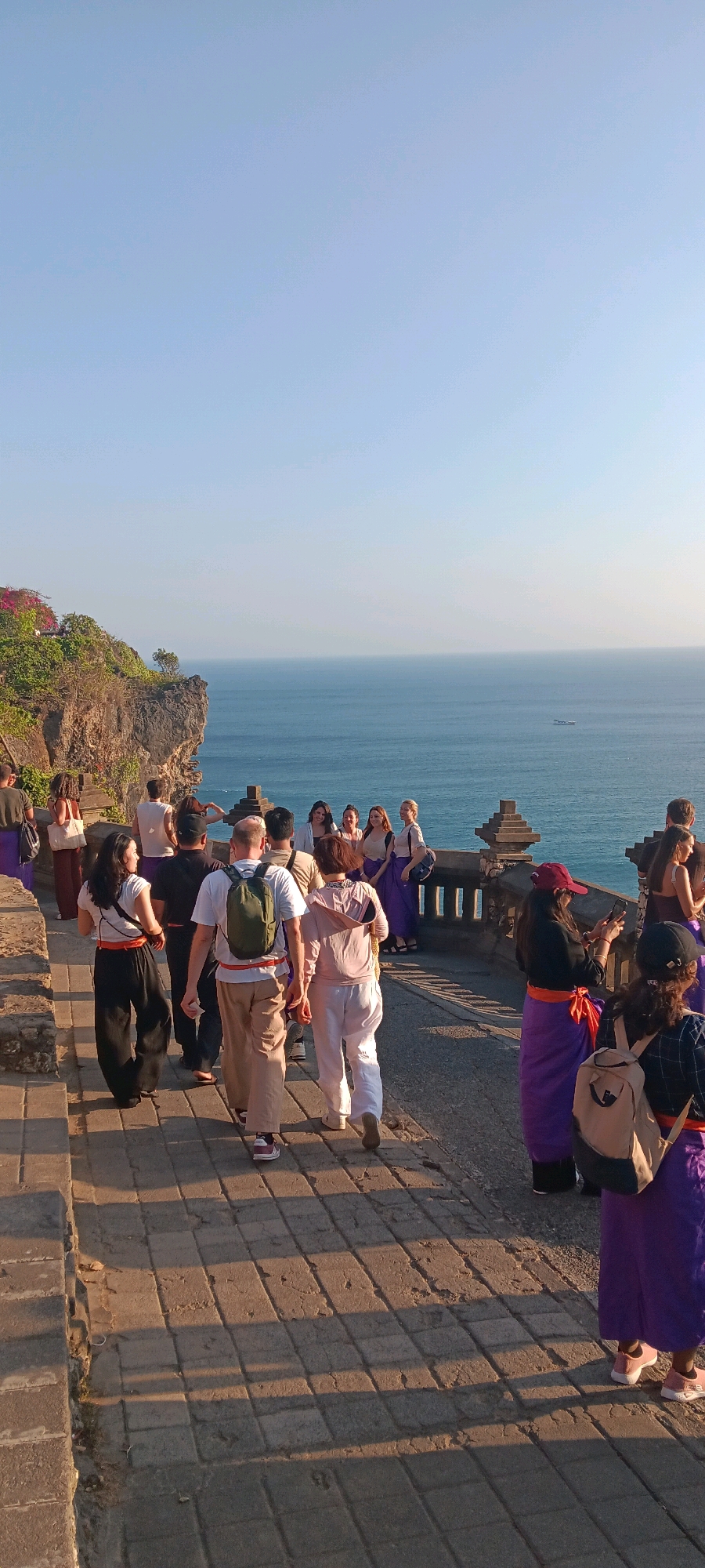 In Kuta Selatan, Indonesia, a group of people are gathered on a cliffside promenade overlooking the ocean. The promenade is paved with stone tiles, and a stone railing with decorative elements lines the edge. Lush green vegetation and a rocky cliff face are visible on the left, with vibrant pink flowers blooming near the top. The vast expanse of the blue ocean stretches to the horizon, where a small boat is discernible. The sky is clear and a pale blue.
The people in the image appear to be a mix of tourists and locals. Many are wearing traditional Indonesian attire, characterized by sarongs or loose-fitting pants in shades of purple and orange, paired with light-colored tops. Several individuals are carrying backpacks, suggesting they are exploring the area. Some are walking along the promenade, while others are paused, looking out at the ocean or interacting with each other. One woman in the foreground is taking a picture with her phone, and a group of people stand together near the railing, posing for a photograph. The setting sun casts long shadows across the paved walkway.