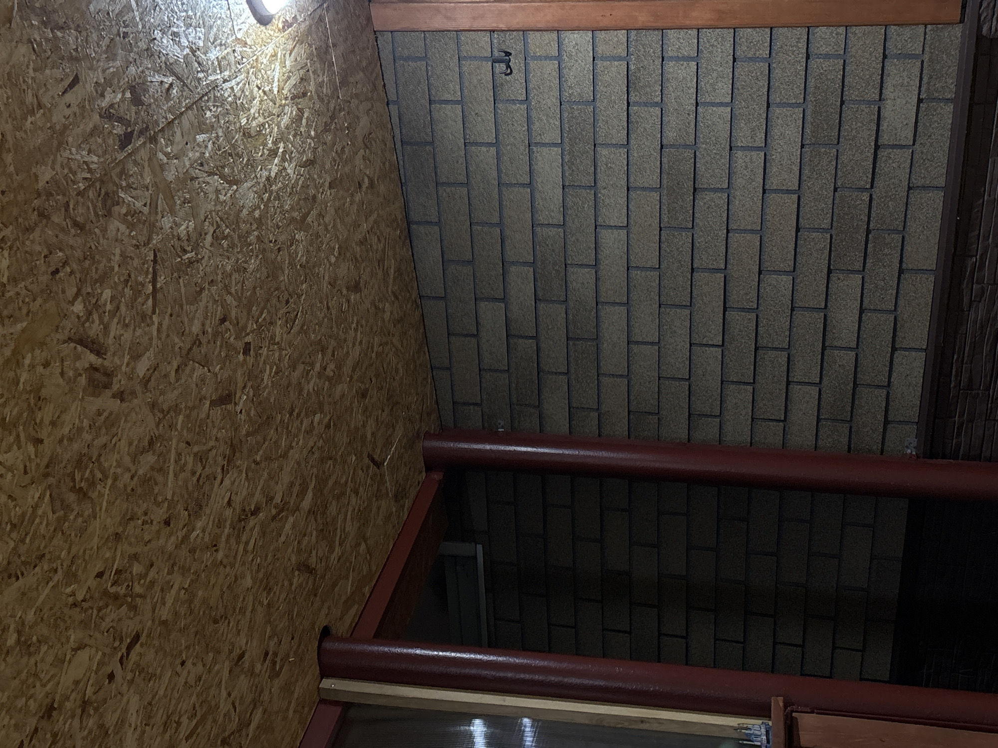 The image depicts an interior space. One wall is constructed of oriented strand board (OSB). The ceiling is covered with rectangular asphalt shingles, with a metal hook attached. In the foreground is a red metal structure, possibly a window frame or part of a bed frame. The light source is a small bulb mounted on the OSB wall. The location is purportedly in Prikamskii, Russia.