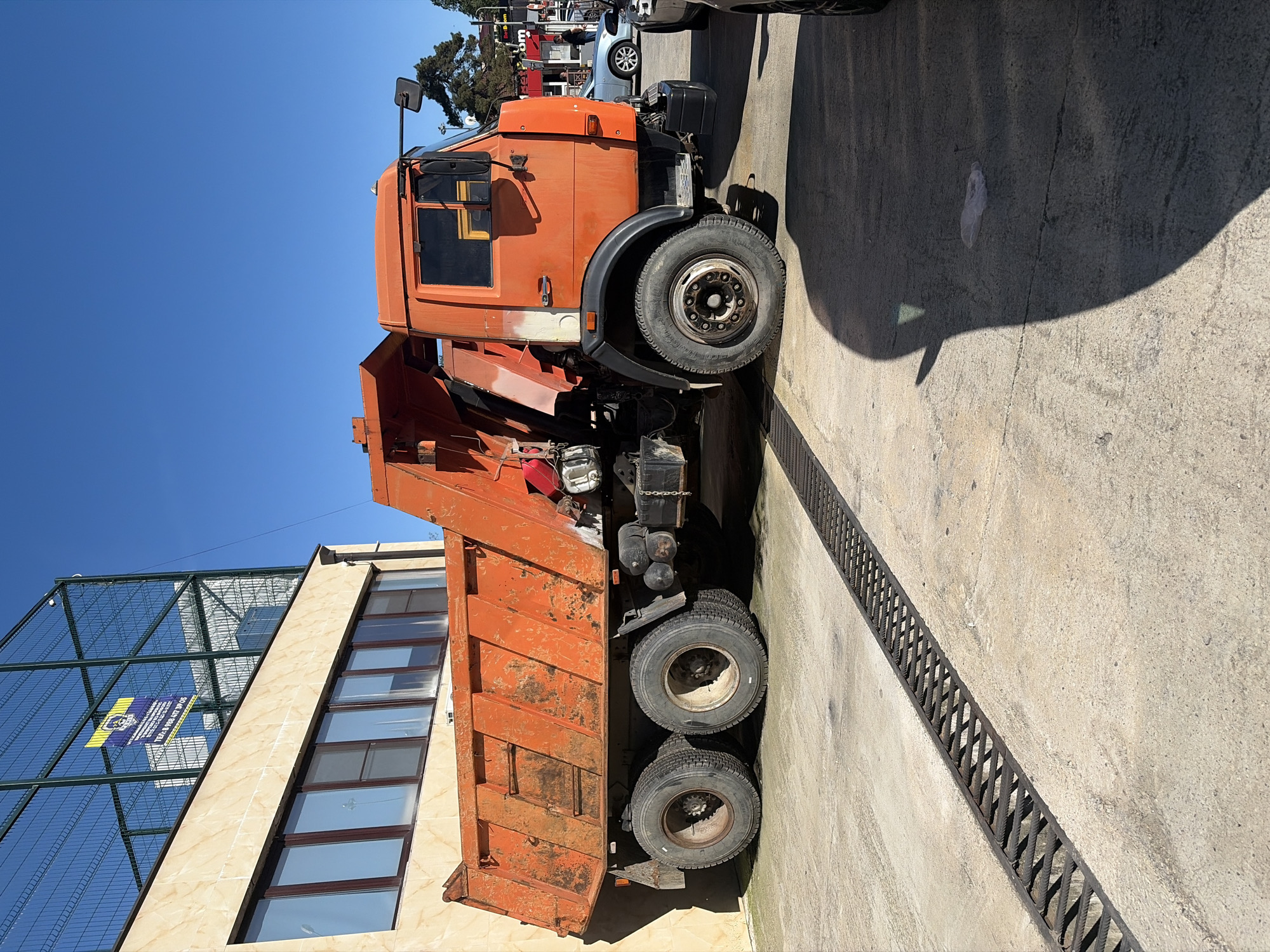 The image features a large, orange dump truck parked outdoors on a concrete surface under a clear blue sky. The truck, an older model showing some wear, is seen in profile, facing towards the right. Its cab, single front wheel, and two sets of rear wheels (dual axles) with their tires are visible, as is the raised dump bed behind the cab.
To the left of the truck stands a multi-story building clad in light-colored tiles, featuring multiple horizontal rectangular windows. Behind this building, a tall, metal mesh fence with square grids is visible, suggesting a sports facility or an industrial enclosure. Attached to this fence is a blue and yellow sign with partially legible text. Visible details include "MEGA...", a phone number "8 928 4** *** 05", and the Russian Ruble symbol (₽), indicating an advertisement likely in a Russian-speaking region. Other vehicles and trees are faintly discernible in the far background.
The ground is a light-colored concrete pavement. A long, dark metal drainage grate runs diagonally across the concrete in the lower and central part of the image. Strong, distinct shadows are cast by the truck and possibly other objects onto the pavement, signifying bright, direct sunlight and clear weather, likely mid-day to early afternoon. The truck is stationary, and no specific activity is taking place. The overall setting appears to be an industrial or commercial parking lot.