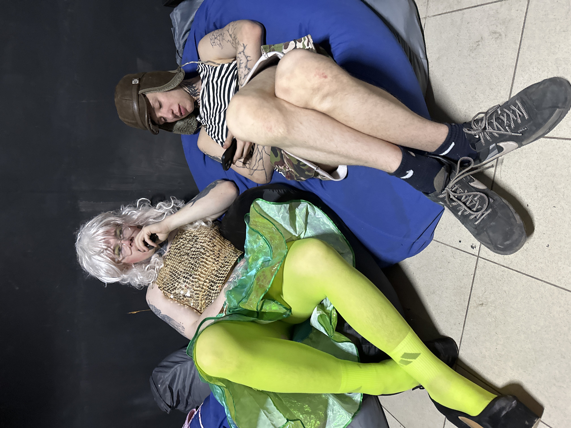 This image, taken indoors in Saratov, Russia, depicts two individuals seated on beanbag chairs against a dark, featureless wall with a tiled floor.
The individual on the left is seated on a dark-colored beanbag, wearing a long, wavy, light-blonde wig and round glasses. Tattoos are visible on both arms and neck. This person is dressed in a gold, sequined strapless top, a layered, ruffled bright green skirt, neon green opaque tights, and black high-heeled shoes. The right hand holds a dark, cylindrical object, potentially a vape pen, near the mouth. Legs are crossed at the ankles.
The individual on the right is seated on a blue beanbag, wearing a brown aviator-style hat with ear flaps, a black and white horizontally striped tank top, and camouflage shorts. Extensive tattoos cover both arms and the neck. This person holds a black smartphone with both hands, looking downwards at the device. Legs are crossed at the knees. The individual wears dark socks with white stripes and dark sneakers with white laces, labeled "UNILA" on one shoe. A visible abrasion is present on the right knee.
The floor consists of light-colored square tiles with dark grout lines, showing some marks and wear. A crumpled dark grey object, possibly another beanbag or fabric, is visible behind the left individual. No direct interaction between the two individuals is apparent.