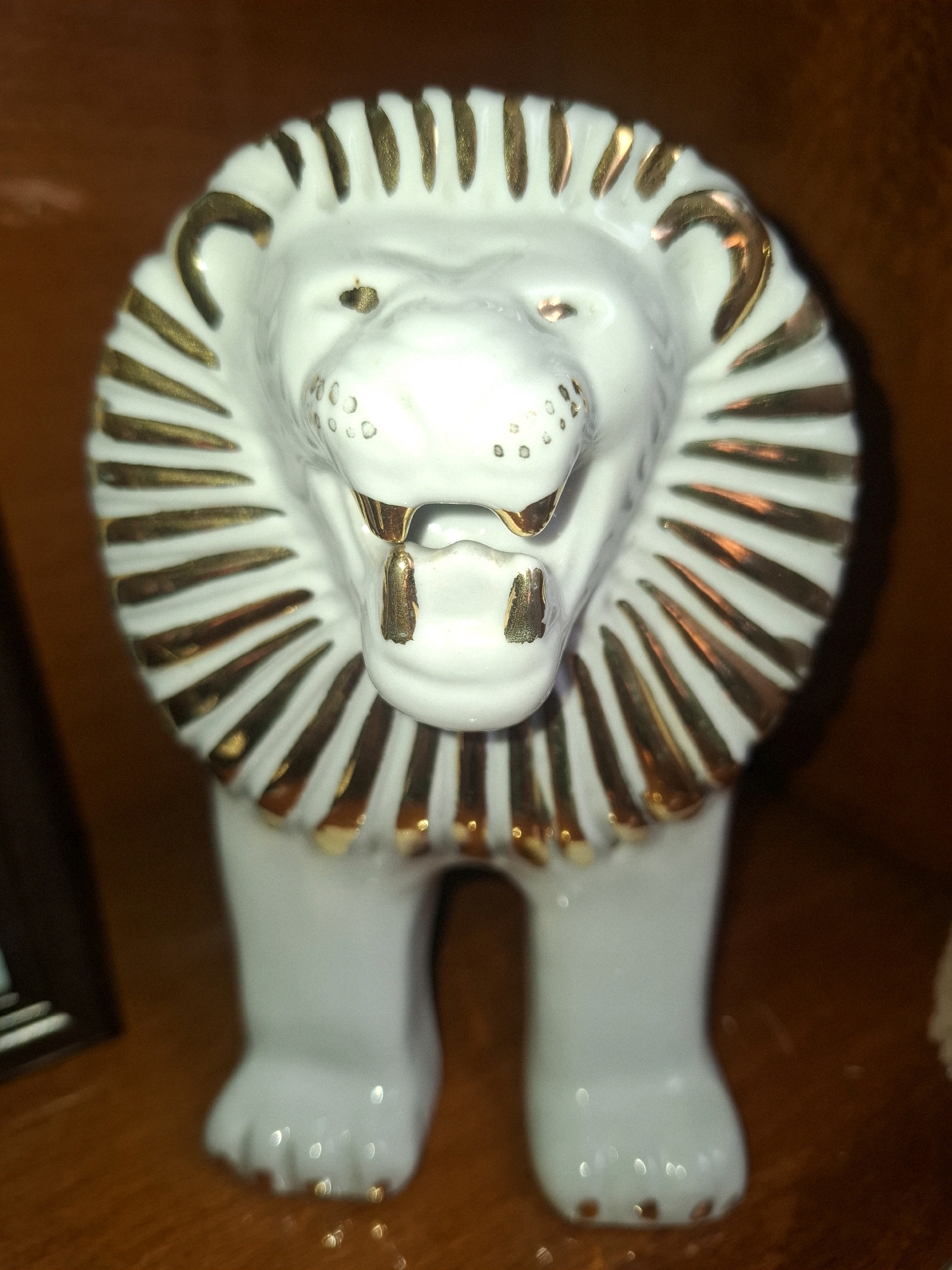 A close-up, eye-level studio photograph shows a white ceramic lion figurine with gold accents. The figurine is depicted from the chest up, facing the camera. Its mane is represented by radiating gold-trimmed segments, and its face features gold-outlined eyes, nostrils, and an open mouth with visible gold fangs and tongue. Small gold dots suggest whiskers or facial texture. The lion is standing on all four paws, though only the front legs are clearly visible. The figurine is placed on a warm brown, wooden surface, likely a shelf or table. The background is a blurry, dark brown, suggesting a dimly lit indoor environment. The lighting highlights the shiny surfaces of the ceramic and gold elements. There is no discernible location in the city of Almaty, Kazakhstan; this appears to be a staged product photograph or a personal item display.