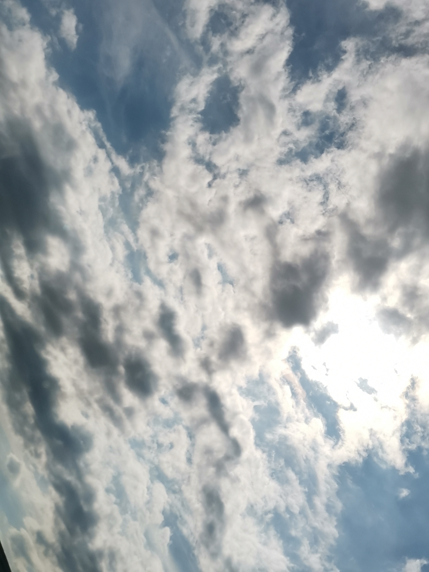The image is a photograph of a sky filled with a mix of white and dark gray clouds against a blue sky. The sun is partially obscured by the clouds, creating a bright, washed-out area in the upper right portion of the image. The clouds are varied in shape and density, with some appearing wispy and others more solid and textured. The image does not contain any discernable elements that would allow for the identification of a specific location within the city of Sofia, Bulgaria, or any people, subjects, objects, or actions. The scene is purely natural, depicting a cloudy sky.