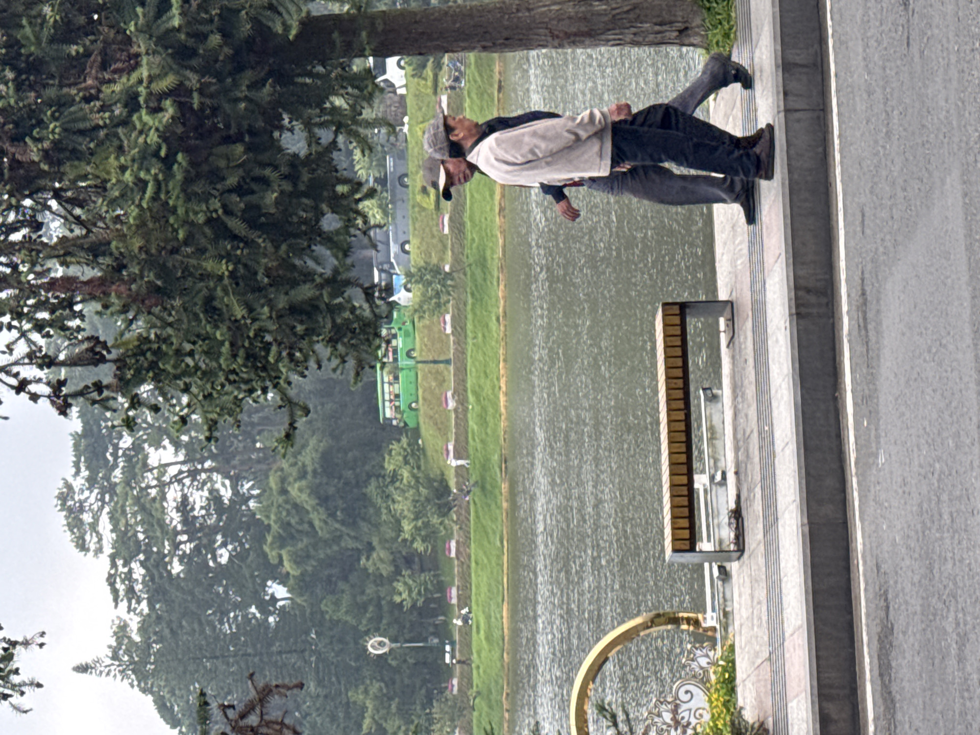 Two middle-aged men are walking along a paved walkway beside a lake. The man on the left is wearing a light-colored jacket and dark pants, while the man on the right is wearing a dark jacket and dark pants. They are both facing to the right and appear to be in conversation. In the background, there is a body of water, a grassy embankment with trees, and several buses parked on a road. A bench is visible on the walkway. The overall scene suggests a public park or recreational area in Da Lat, Vietnam, likely Xuan Huong Lake, a popular tourist destination known for its scenic views and surrounding greenery. The weather appears overcast.