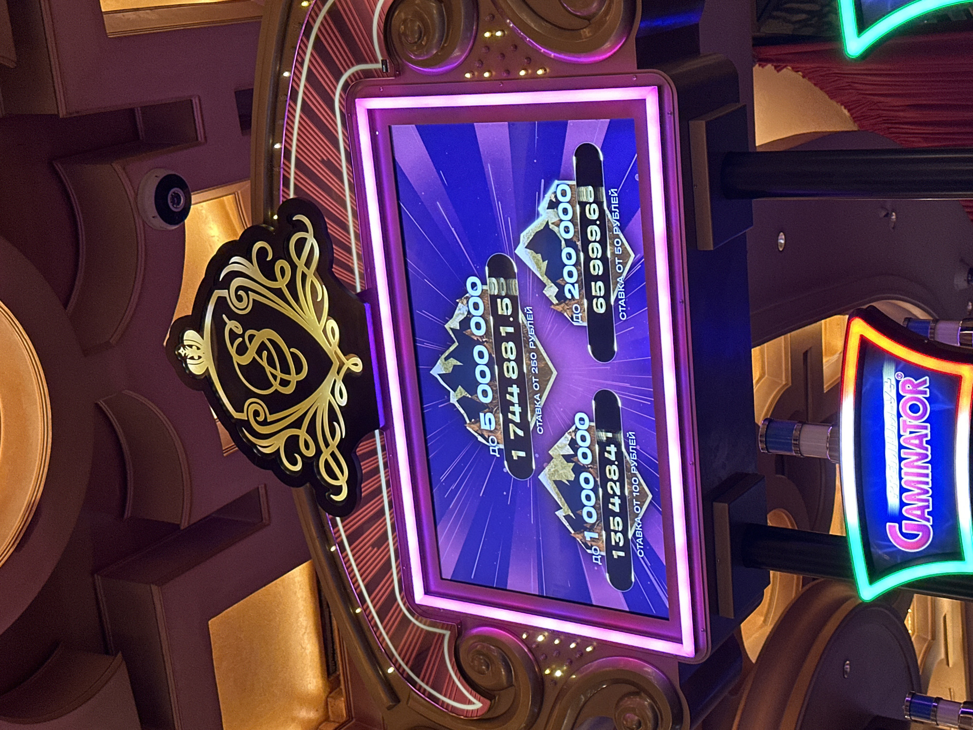This image shows the interior of a casino in Estosadok, Russia, specifically a Gaminator slot machine. The slot machine has a large, illuminated screen displaying a progressive jackpot system. The screen features mountains and numbers indicating different jackpot tiers: "до 5 000 000" (up to 5,000,000) with a current value of "1 744 881.5", and "до 1 000 000" (up to 1,000,000) with a current value of "135 428.41". Below these are indications of minimum bets: "СТАВКА ОТ 250 РУБЛЕЙ" (bet from 250 rubles) and "СТАВКА ОТ 100 РУБЛЕЙ" (bet from 100 rubles). To the right, another jackpot tier is visible: "до 200 000" (up to 200,000) with a current value of "65 999.65" and a minimum bet of "СТАВКА ОТ 50 РУБЛЕЙ" (bet from 50 rubles). The machine itself is adorned with ornate golden detailing and purple lighting. The Gaminator logo is prominently displayed at the bottom of the screen with neon green and red lighting. The ceiling of the casino is visible in the background, featuring intricate architectural details and lighting. A security camera is mounted on the ceiling. No people are visible in the image.