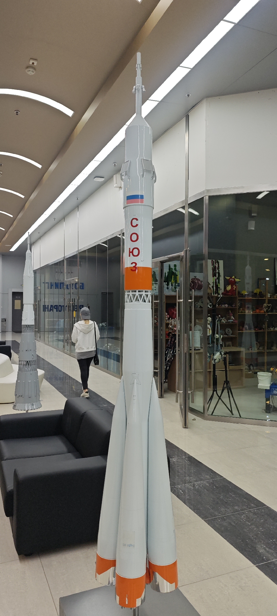 The image shows the interior of a building in Estosadok, Russia. Two models of the Soyuz rocket are prominent; the larger model is painted white, orange, and grey, with the Russian flag and the Cyrillic letters "COЮ3" painted on its side. The second model, smaller and grey, stands in the background. A woman in a white hooded sweatshirt and dark pants is walking past a black sofa towards the background. The foreground exhibits a tiled floor that features a black and white patterned design. The background includes a glass-walled store displaying various items, including toys and equipment. The ceiling has recessed lighting, which casts illumination throughout the area.
