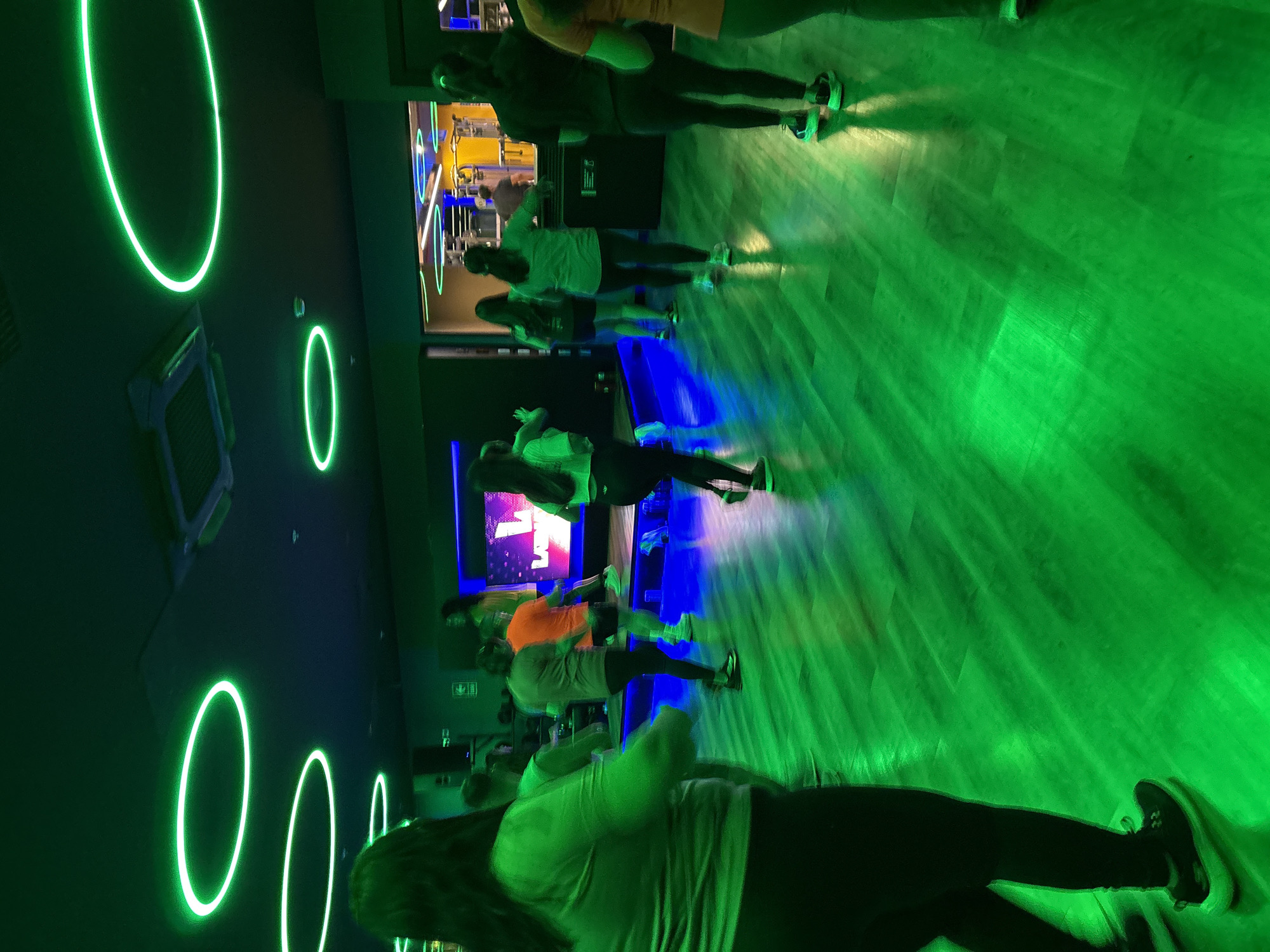 The image depicts a group of people participating in a fitness class, likely a dance or aerobics session, in a dimly lit studio. The room is illuminated with vibrant green neon lights, casting a strong green hue over the scene, particularly on the wooden floor. Circular neon lights are also visible on the dark ceiling.
In the foreground, the back of a woman wearing a light-colored t-shirt and black leggings is prominent, with her dark hair flowing. She appears to be in motion, suggesting she is actively engaged in the class. Several other individuals are visible further back, also in various stages of movement. One person in the mid-ground wears an orange t-shirt. Behind the participants, a large screen displays the word "LATIN" in large, colorful letters. The floor is made of wooden planks that reflect the green light.
In the background, through an opening, a section of a gym or fitness equipment area is visible, with yellow and metallic structures suggesting exercise machines. The overall atmosphere is energetic and modern, consistent with a contemporary fitness studio. The location is identified as San Isidro, Peru, a district known for its upscale businesses and residential areas.
