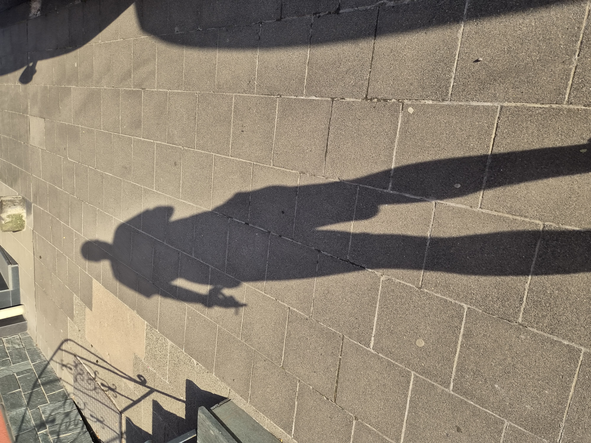 A shadow of a person stands on a paved surface. The shadow is long, indicating the sun is low in the sky. The paving stones are rectangular and laid in a pattern. There is a railing and steps to the left of the shadow. The area appears to be an outdoor public space, possibly a street or pathway. The image does not provide any specific information about the location within the city of Sanem, Luxembourg.