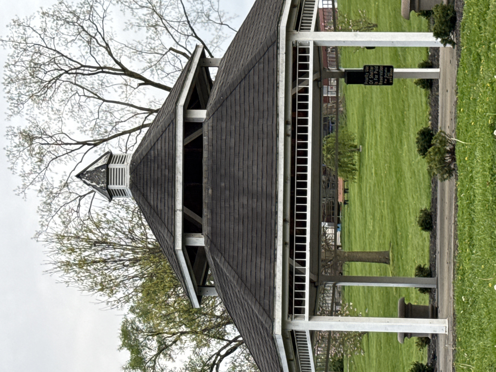 The image depicts a gazebo located in Grove City, Ohio, United States of America. The gazebo has a multi-tiered roof with dark gray shingles. White posts support the roof structure. The gazebo is surrounded by a lawn with green grass. A sign is visible that says "Priority Use Of Shelter Is For Paid Reservation. For Details 277-3000". Some shrubs are planted next to the gazebo. Trees with bare branches are visible next to the structure. A cloudy sky is overhead.