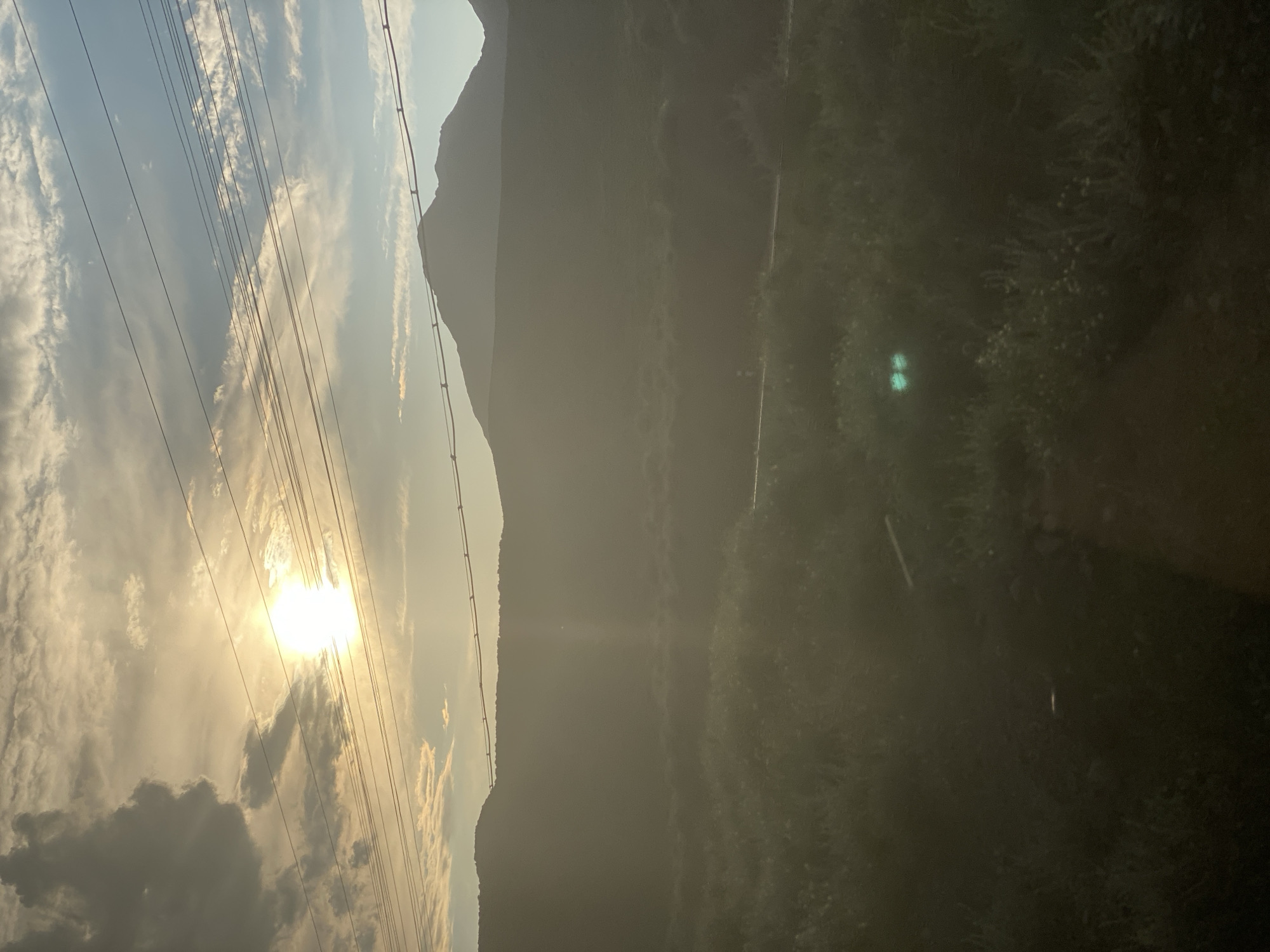 This is a medium shot photograph of a mountainous landscape under a hazy, sunlit sky. The sun is partially obscured by clouds and appears to be setting or rising, casting a warm, diffused light across the scene. Overhead, several power lines stretch horizontally across the frame. In the middle ground, rolling hills are silhouetted against the sky, and the foreground consists of dense vegetation, possibly shrubs and grasses, with some small white flowers visible. Two bright, glowing green circles are present in the vegetation, likely the reflection of light from an unknown source. The overall impression is one of a natural, rural environment within the vicinity of Kok-Moinok - 1, Kyrgyzstan.