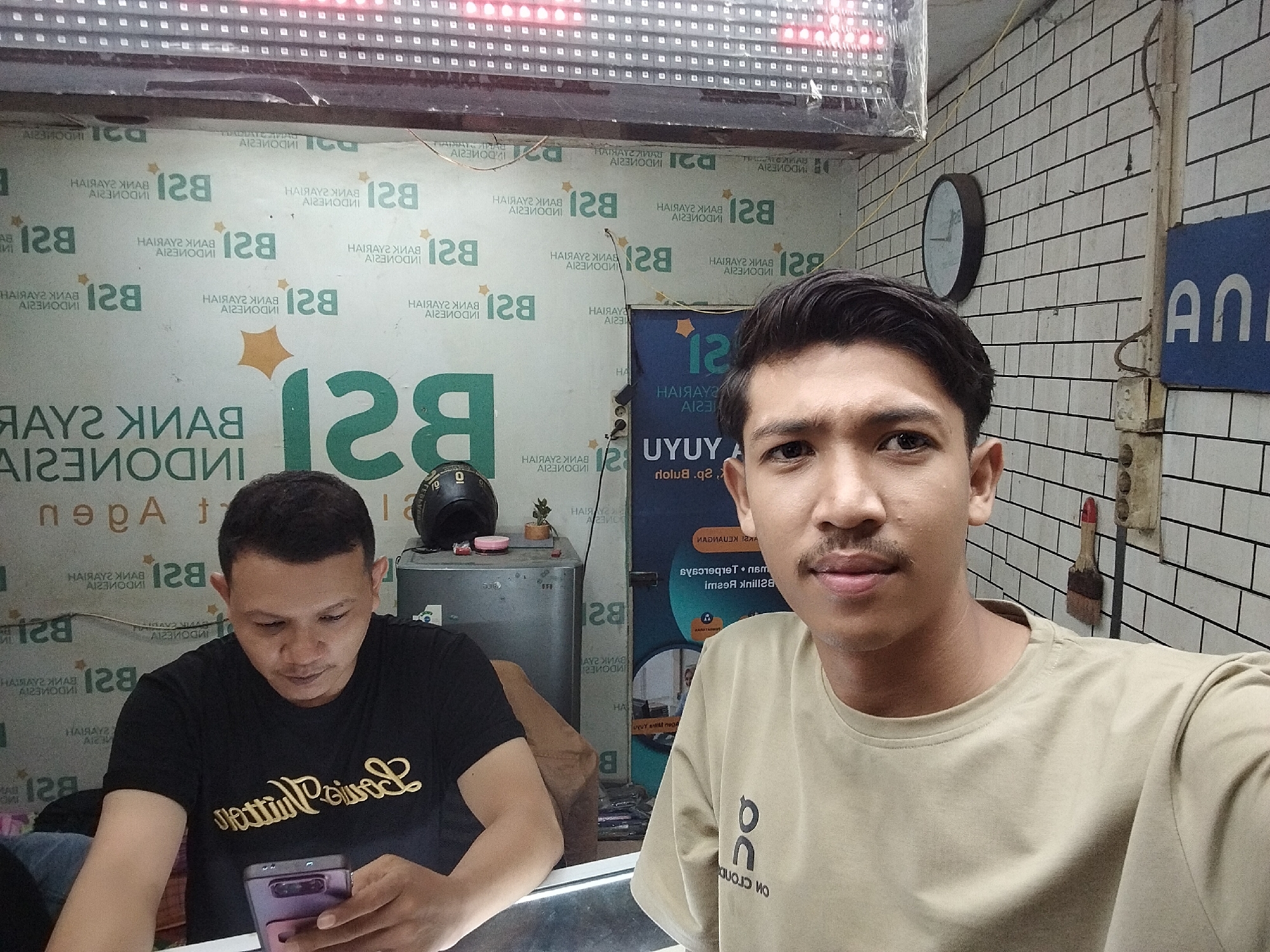 Two men are captured indoors, likely in an agent office for Bank Syariah Indonesia, located in Lhokseumawe, Indonesia. In the foreground, a man wearing a beige t-shirt with an "OC" logo is taking a selfie, looking directly at the camera with a neutral expression. Behind him and to the left, another man in a black t-shirt, which appears to have "Letture Private" or similar script, is looking down and focused on a purple smartphone he holds.
The background is busy with branding. The wall behind the men is covered with repeated logos and text of "BSI" and "BANK SYARIAH INDONESIA", many of which are mirrored. A partially visible blue banner or poster behind the man in beige also shows text like "UYUY" and "UYUYA". To the left, a small refrigerator or cabinet holds a black helmet and a small potted plant. The ceiling features an illuminated LED display with red lights. To the right, a wall of white subway tiles features a round wall clock and an electrical outlet. A paintbrush is hanging near the outlet. The overall setting suggests a casual, possibly commercial, indoor environment.