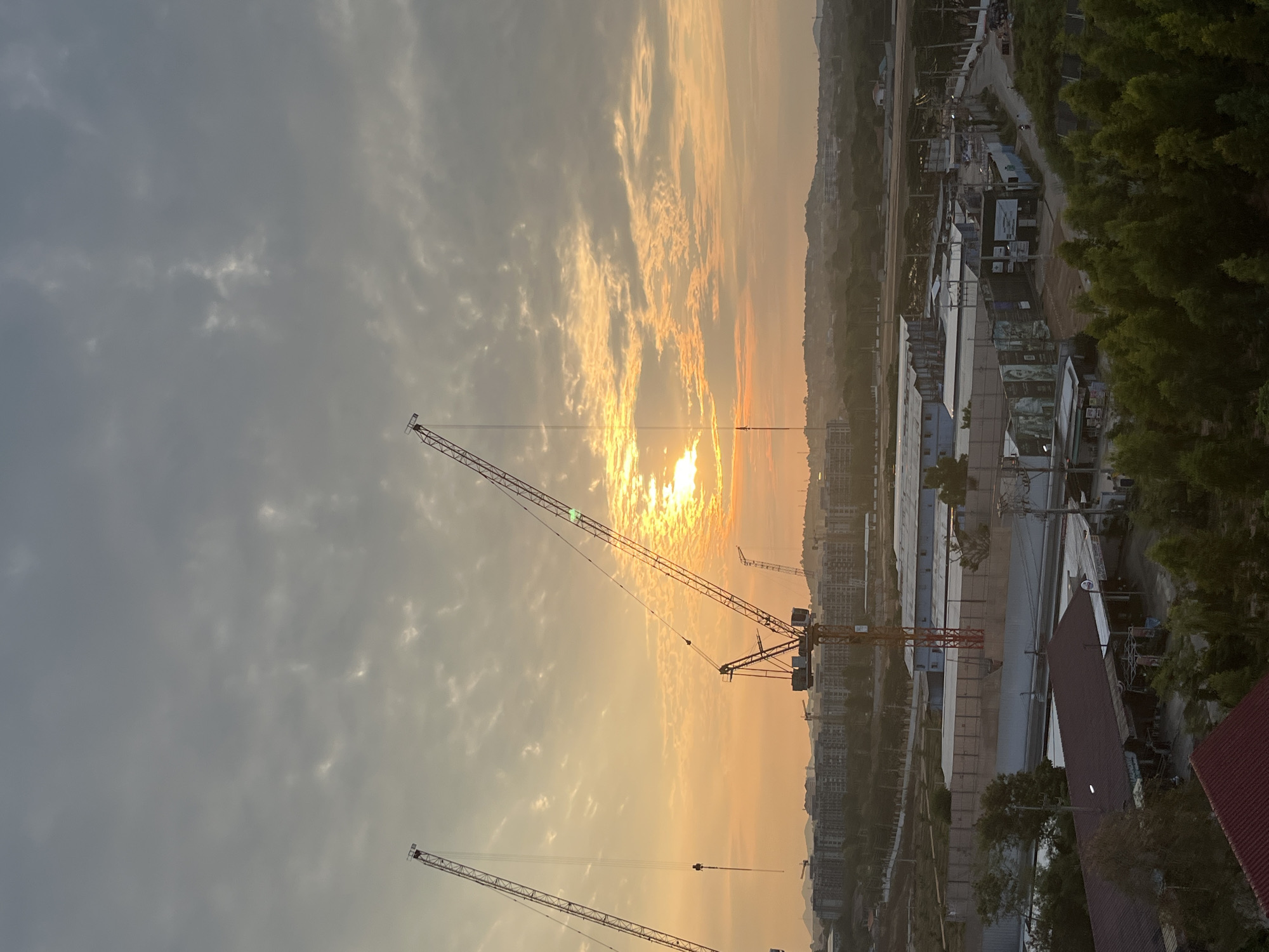 The image captures a cityscape within Pattaya City, Thailand, at either sunrise or sunset. The sky is dominated by a mix of clouds and a strong, diffused sunlight source creating a golden hue. Several construction cranes are silhouetted against the light, suggesting ongoing construction activities. Below, there is a blend of residential and industrial structures, including building roofs, warehouses, and green vegetation along the edges. The perspective indicates that the shot was taken from an elevated position, possibly a high-rise building or a hilltop.
