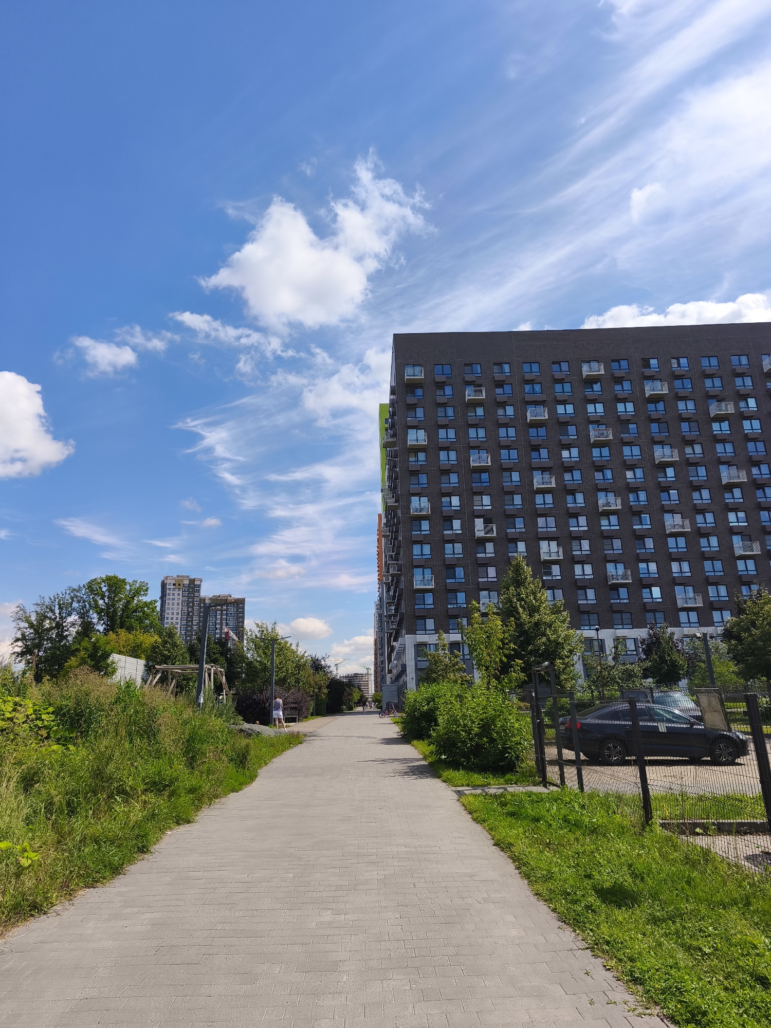 The image depicts a modern residential area on a sunny day, likely in Moscow, Russia, given the architectural style and vegetation. A paved walkway, made of grey rectangular stones, stretches into the distance, bordered by lush green grass and bushes on the left and a metal fence with more greenery and parked cars on the right. Prominently featured is a large, dark-colored, multi-story apartment building with numerous windows and small balconies, dominating the right side of the frame. In the background, other tall buildings are visible, suggesting a dense urban environment. The sky is a vibrant blue, dotted with scattered white clouds, some appearing as wispy cirrus formations. A few trees with green foliage are interspersed throughout the scene. A person, dressed in light-colored shorts and a grey top, is visible walking away from the camera on the path. Further down the path, another individual appears to be on a bicycle. The overall impression is one of a developed urban neighborhood with green spaces, characteristic of contemporary Moscow housing developments.