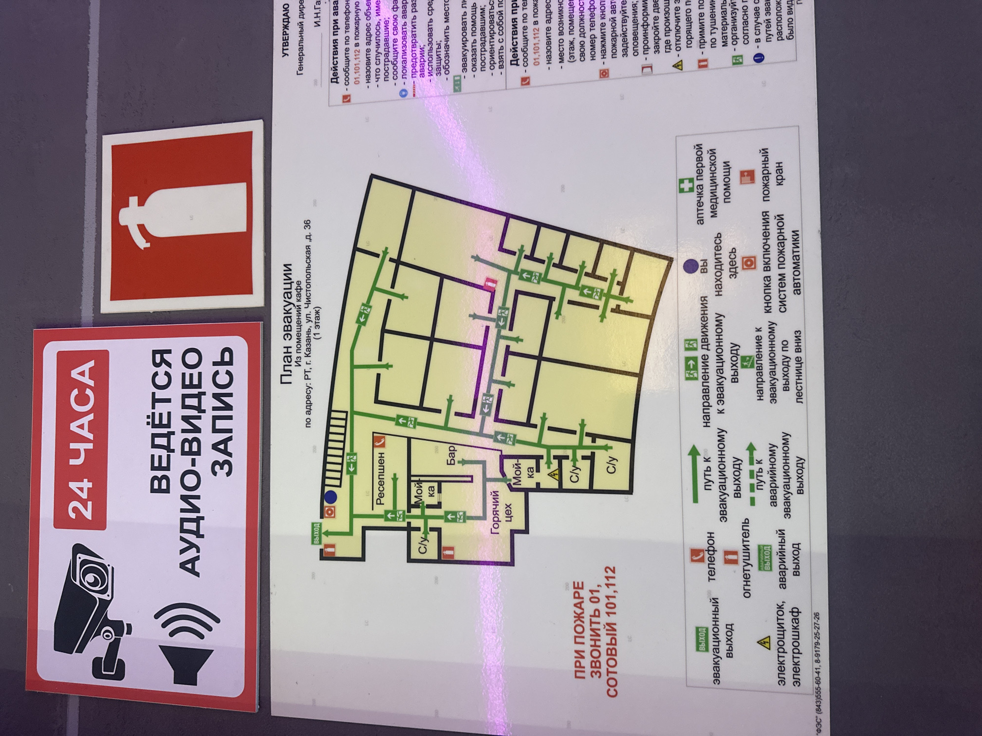 The image shows a sign for an evacuation plan from a cafe in Kazan, Russia. The sign includes a map of the cafe, showing the location of exits, restrooms, a bar, and a hot kitchen. There are also instructions on what to do in case of a fire, including the phone numbers to call. Next to the evacuation plan is a sign indicating that audio and video recording is in progress 24 hours a day. A fire extinguisher sign is present.