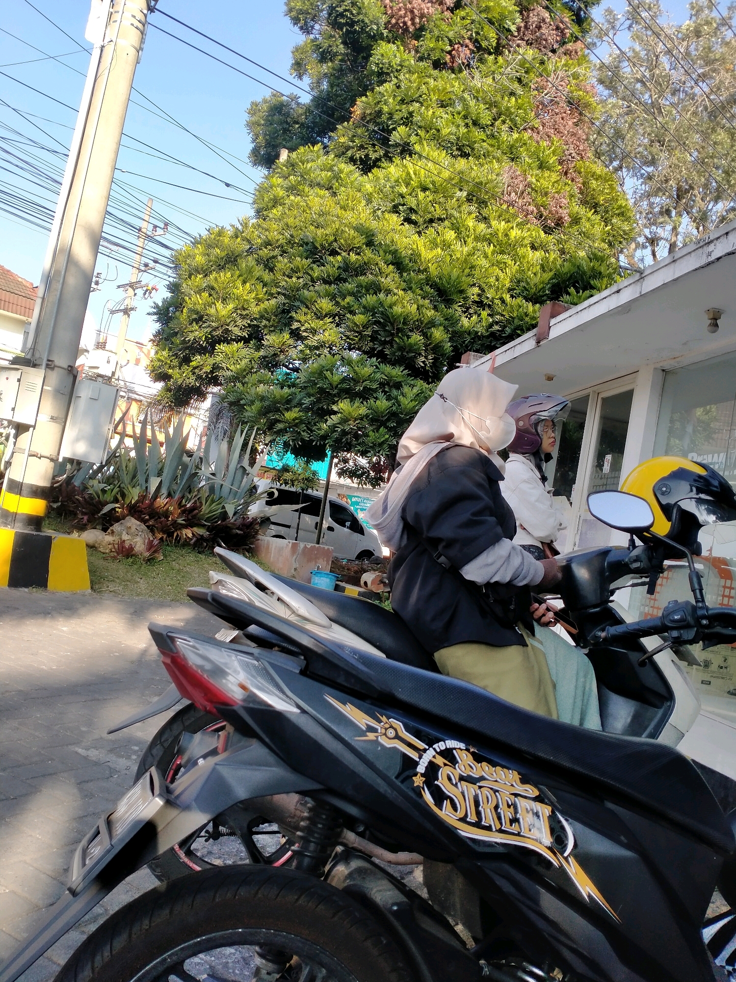 The image depicts a street scene in Batu, Indonesia, with a focus on two individuals on a motorcycle. A woman wearing a light-colored hijab and a dark jacket, and green pants, is seated on the front of the motorcycle. She is wearing a white face mask. Behind her, another person wearing a helmet sits on the back of the motorcycle. The motorcycle itself is black with yellow and white decals on the side that read "Beat Street" and "Born to Ride". A yellow helmet is perched on the handlebars.
The environment includes a paved street with yellow and black striped bollards on the left side. A large tree with lush green foliage is prominent in the background, casting shadows. Adjacent to the tree is a white building with a window and a light fixture. Power lines crisscross the sky, supported by a tall utility pole. A patch of ground with various plants, including spiky agaves and reddish bromeliads, is visible between the bollards and the building. A white van is parked in the background. The sky is clear and blue, indicating a sunny day.