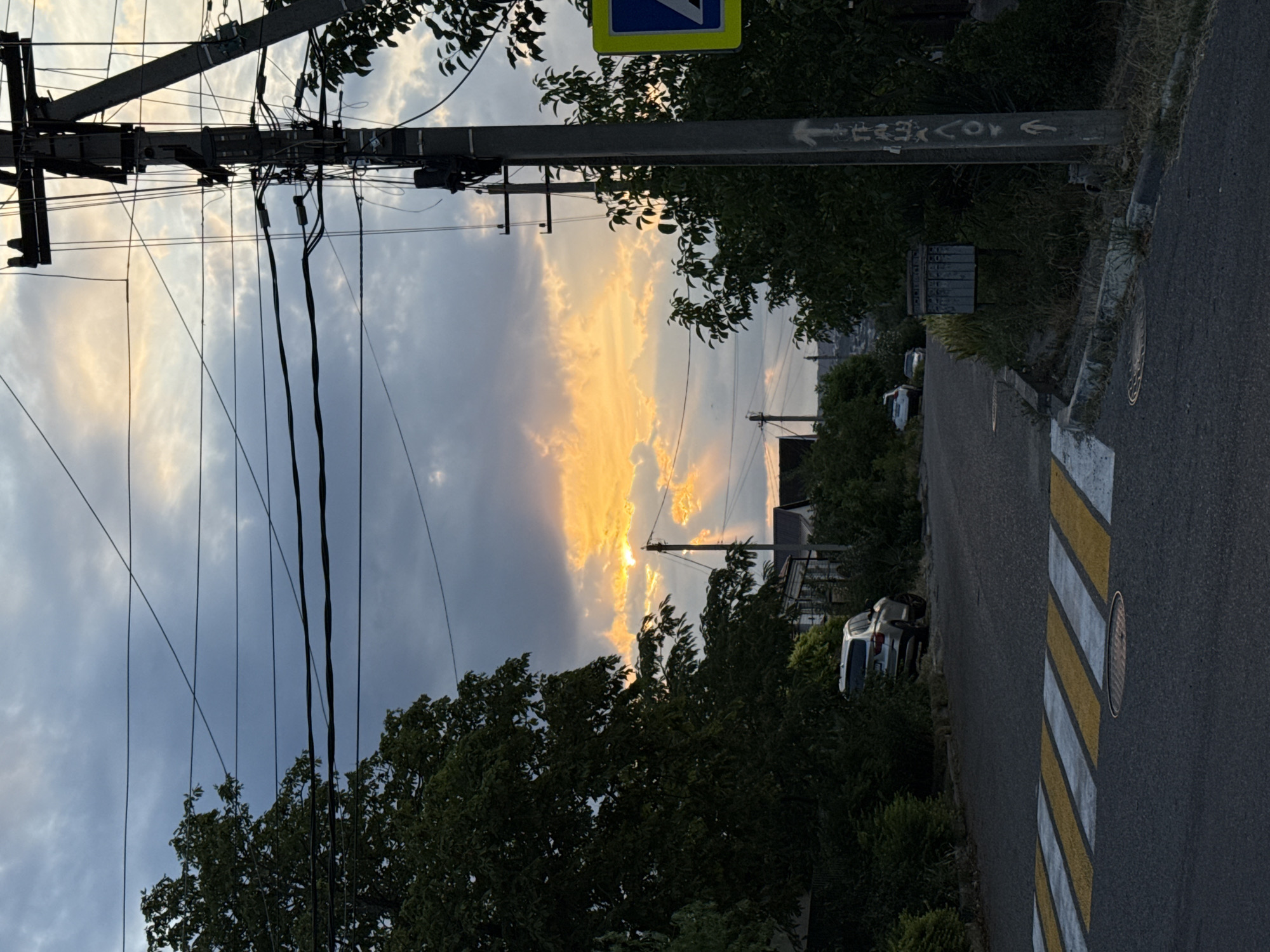 This image depicts a street scene in Novorossiysk, Russia, likely during sunset or sunrise. A prominent power pole with numerous wires dominates the right side of the frame, stretching towards the sky. The sky itself is dramatic, with heavy clouds partially obscuring a bright, golden sun that casts rays of light through the openings.
In the foreground, a pedestrian crossing with yellow and white stripes is clearly visible on the asphalt road. A storm drain cover is located near the crossing. On the left side of the road, a mature tree with lush green foliage overhangs the street. Further down the road, a white SUV is parked on the side, partially obscured by vegetation. Several houses with white roofs can be seen in the background, nestled amongst trees and hills.
A blue and yellow traffic sign is attached to the power pole on the right. Adjacent to the pole, a metal mailbox unit with multiple compartments is situated amidst overgrown grass. The overall impression is of a residential street bathed in the dramatic light of a setting or rising sun, with the functional elements of urban infrastructure present.