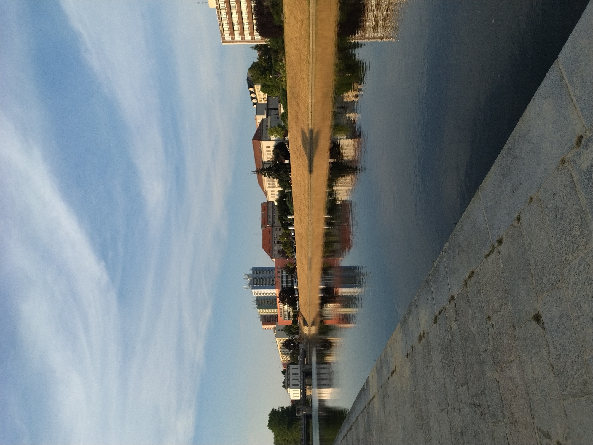 The image depicts a wide, calm river flowing through a city, likely Písek, Czechia, given the architectural style and the presence of a distinct bridge structure. The foreground shows a paved walkway made of large, grey stone slabs, with small tufts of grass growing between them. This walkway extends along the riverbank, suggesting a promenade or quay.
The river itself is dark and reflects the sky and the buildings on the opposite bank. The water is still, with only faint ripples visible. The opposite bank is lined with a steep, grassy embankment, dry and golden in color, likely due to the season or time of day.
Beyond the embankment, a series of buildings are visible. These include several multi-story residential buildings with colorful facades (orange, yellow, light blue), and some older, lower-rise buildings with red-tiled roofs. One prominent building on the far left appears to be a white, symmetrical structure with large windows, possibly a public building or institution, with a bridge extending over the river in front of it. Further down the river, a modern, tall building with many windows is also visible.
The sky above is a clear blue, with scattered, wispy white clouds that catch the sunlight. The overall atmosphere is tranquil and serene, suggesting a peaceful urban scene, possibly during the late afternoon or early morning. There are no people visible in the image.