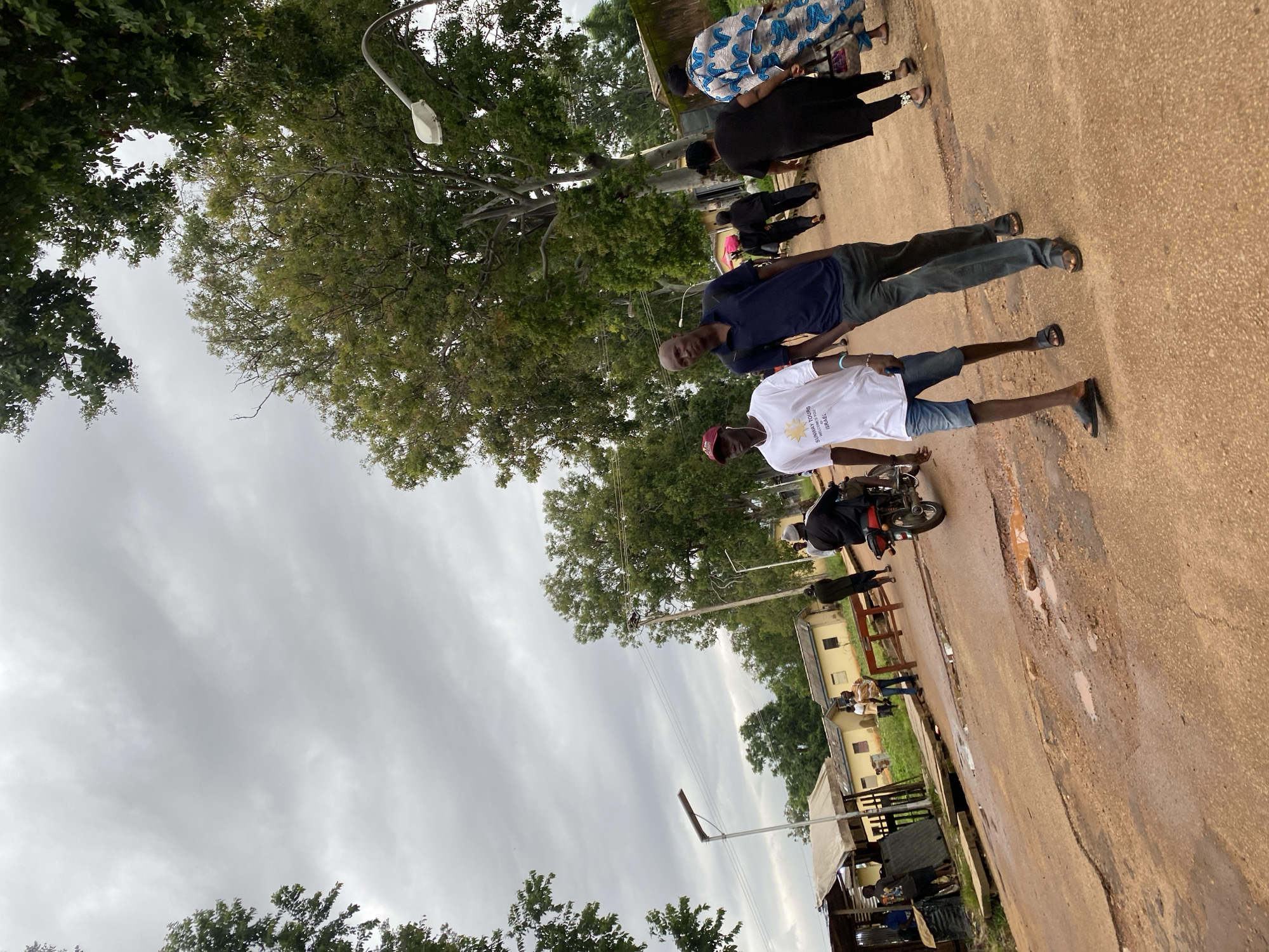 The image captures a street scene in Rafin Par, Nigeria, on an overcast day. Two men are walking towards the foreground on a muddy, potholed dirt road. The man on the left wears a white t-shirt with text that appears to say "SUNWAY TOURS WELCOMES YOU TO ISRAEL" and blue denim shorts. He has a red baseball cap on. The man to his right wears a dark blue polo shirt and dark pants. Several other individuals are visible in the background. A man rides a motorcycle in the far right, while a group of people, including two women in dark clothing and one in a patterned dress, walk away from the camera. Several low-rise buildings with yellowish walls and corrugated metal roofs are situated along the side of the road, some with awnings. Green trees with dense foliage line the road and a street lamp is visible. The overall environment suggests a somewhat rural or semi-urban setting within the city.