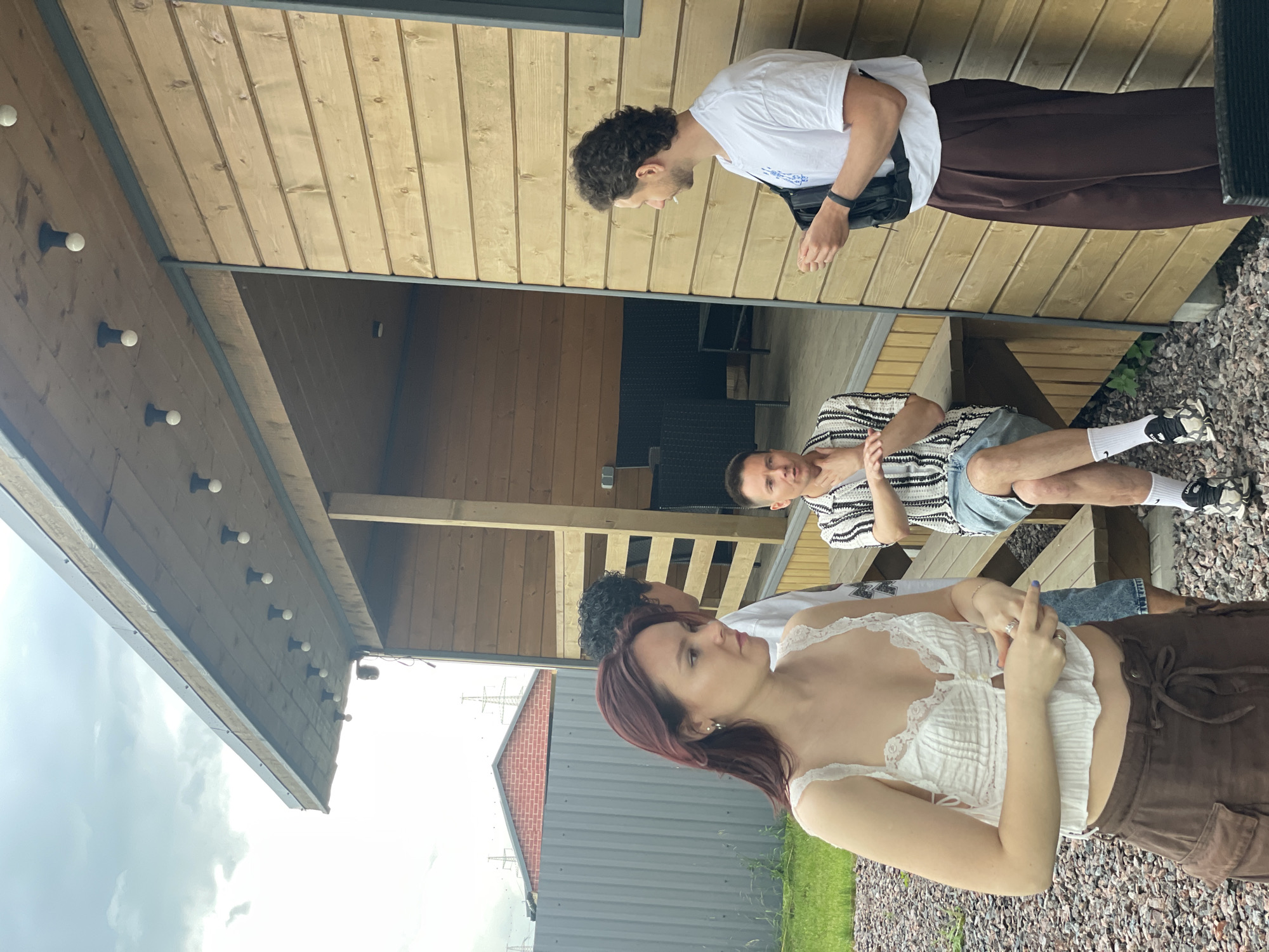 In Novo-Tuutari, Russia, a group of four individuals are gathered outdoors near a wooden building. On the left, a young woman with long, reddish-brown hair is wearing a white lacy crop top and brown cargo pants, facing to her right. To her right and slightly behind her, a man with curly dark hair is partially visible, wearing a white t-shirt and dark pants. In the center, a man with short brown hair is seated on a wooden bench, wearing a striped short-sleeved shirt and blue shorts. On the right, a man with curly dark hair is standing and talking, wearing a white t-shirt with a graphic print and dark brown trousers. He is holding a black object in his left hand. The building behind them is constructed of horizontal wooden planks. The area is covered in gravel, with patches of green grass. Overhead, the sky is cloudy with visible power lines in the distance.
