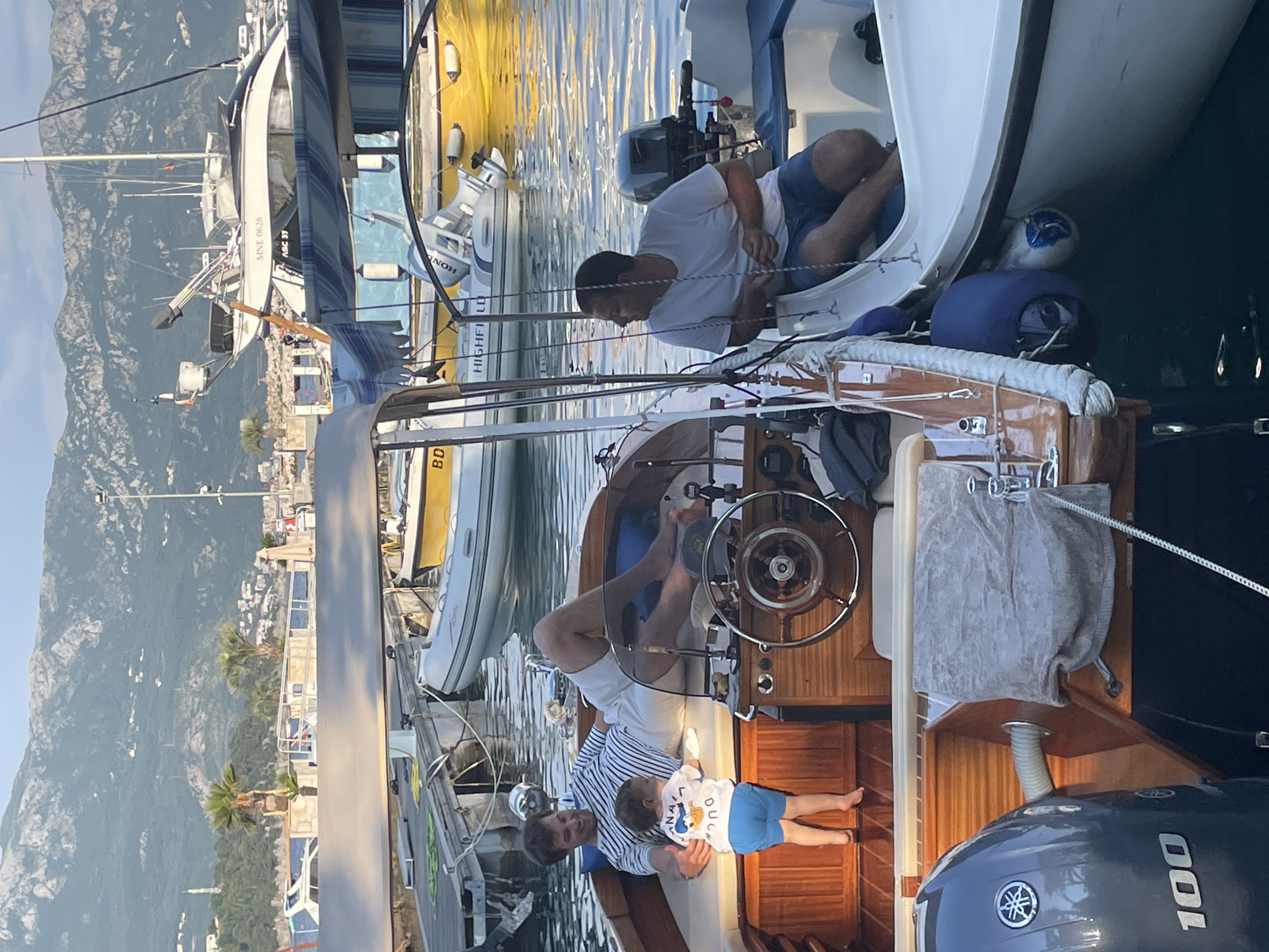 The image shows a marina setting in Budva, Montenegro, with boats docked. Two men are aboard one boat, one sitting and working on something near the bow, the other standing and holding a small child who is lying down. The boat has a wooden interior and a steering wheel. Other boats are visible in the background, along with the city and mountains in the distance.