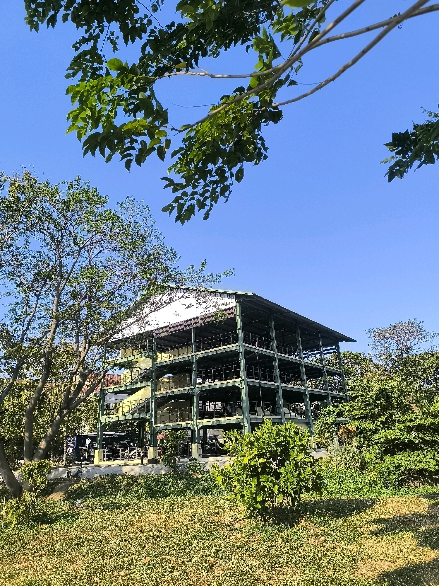 The image captures an outdoor scene with a multi-story structure resembling a parking garage or an open-air building, constructed with a steel frame and green horizontal supports. The structure has at least four visible levels, each with railings and open sides, and is topped with a white roof. The building is situated in a lush, green environment, surrounded by trees and vegetation. In the foreground, there is a grassy area with patches of dry grass and shadows cast by overhead foliage. On the ground level of the structure, a black van with graphics and a motorcycle are visible, suggesting it may be a public or commercial space. The sky is clear and blue. The overall impression is of a modern, open-air building integrated into a natural, park-like setting, possibly a parking facility at a leisure or commercial complex. Based on the architectural style and surrounding vegetation, this location is likely in a tropical or subtropical region.
This image is of the National Geopark Gunung Sewu building in Surabaya, Indonesia. The building is a modern, multi-story structure with an open design, intended to serve as a visitor center or information hub for the geopark. The surrounding vegetation and tropical climate are characteristic of Surabaya.