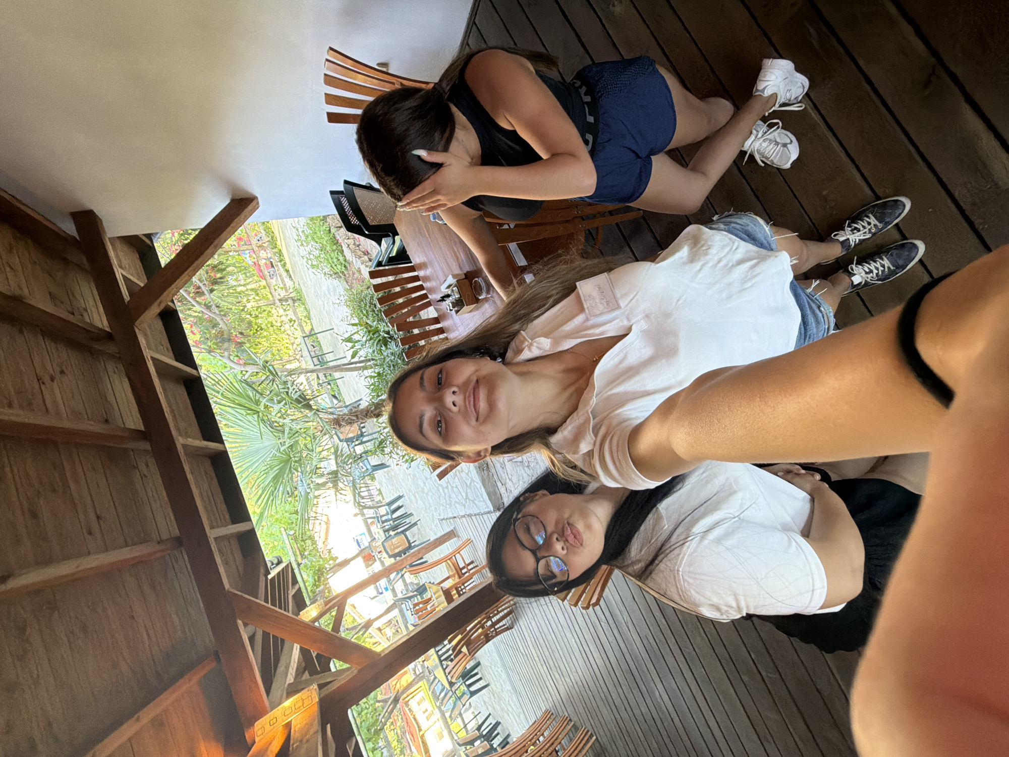 Three young women are photographed from a high angle in an outdoor covered seating area. The woman on the left is wearing glasses and a white t-shirt, and is puckering her lips. The woman in the center is wearing a white collared shirt and denim shorts, and is smiling at the camera. The woman on the right is wearing a black tank top and blue shorts, and has her hand over her face. The area is furnished with wooden tables and chairs, and palm trees are visible in the background. The overall impression is of a casual gathering or outing in a tropical or Mediterranean setting. The location is identified as Tsarevo, Bulgaria.