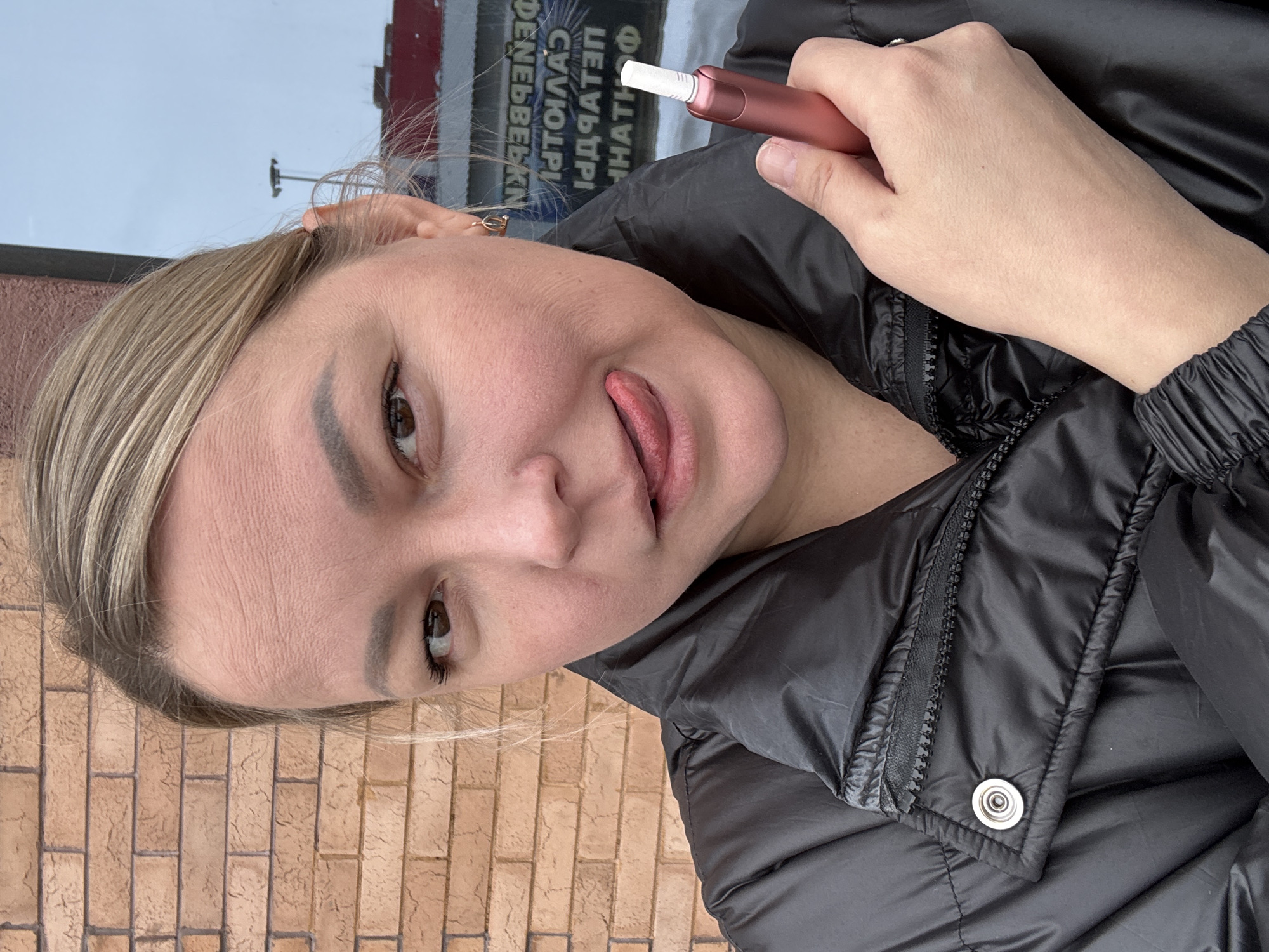 This is a close-up, portrait-style photograph of a woman with a playful expression. The image is framed to show her head, shoulders, and a portion of her arm and hand.
The woman has light-colored hair, possibly blonde or light brown, which appears to be pulled back, with some strands framing her face. She has fair skin, prominent, well-defined eyebrows, and dark brown eyes. She is looking directly at the camera with a lighthearted expression, sticking her tongue out slightly. Visible fine lines and wrinkles are present around her eyes and forehead. She is wearing a black puffer jacket or coat, indicated by the visible textured material, a black zipper running down the front, and a silver snap button closure below the zipper.
In her right hand, which is held up near her face, she is holding a slender electronic device. The device is two-toned, with a metallic rose gold or pink body and a white cylindrical tip, consistent with a heat-not-burn tobacco product.
The background is divided into two main areas. To the left side of the frame, behind her head and shoulder, there is a large, light-colored surface, which appears to be a reflection in a window. Within this reflection, inverted text in Cyrillic script is visible, suggesting a sign or advertisement outside. Partially decipherable mirrored letters include "Ф", "АТН", "ДРІ", "ДІО", "ЛААГ", and "ЕПЕ". A faint red and white structure is also seen in the reflection. To the right side of the frame, behind her body, there is a wall constructed from light brown or orange bricks, showing clear mortar lines.
The lighting suggests it is daytime. The overall mood of the image is casual and candid.