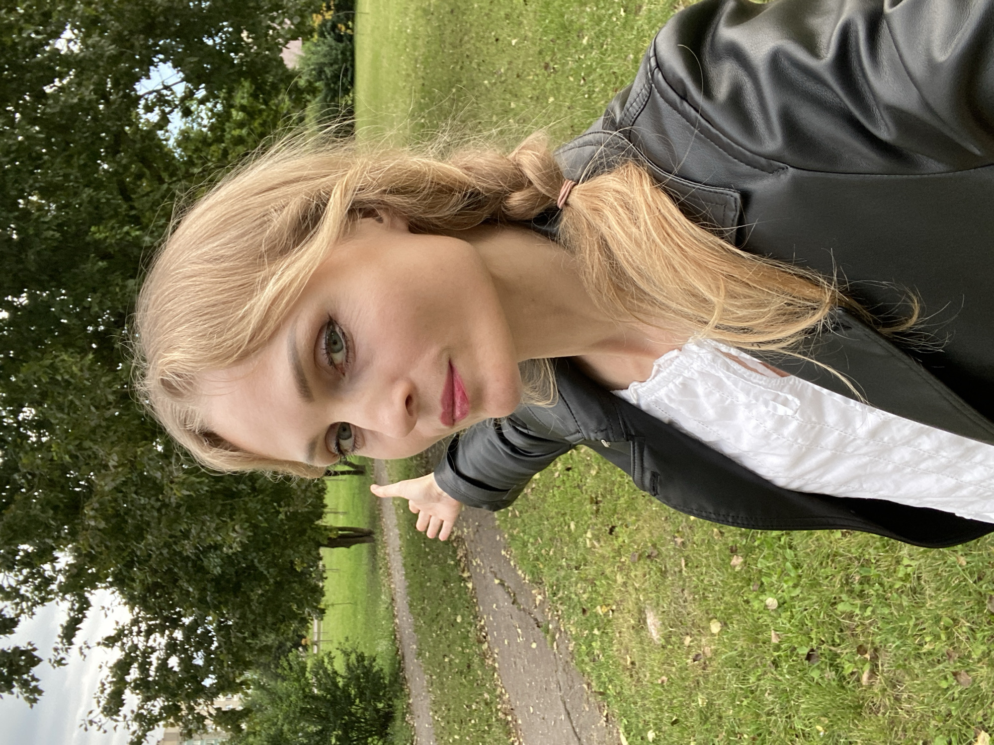 A fair-skinned woman with blonde hair in a braid and wearing a black leather jacket and a white ruffled blouse is captured in a selfie outdoors. She is standing in a park-like setting, with a paved pathway visible to her left and a grassy area with scattered fallen leaves to her right. Tall trees with green foliage form the background, and a multi-story building is visible in the distance. The woman is looking towards the camera with her left hand extended outwards, palm facing upwards. The overall lighting suggests an overcast day. The location is identified as a park in Lida, Belarus.