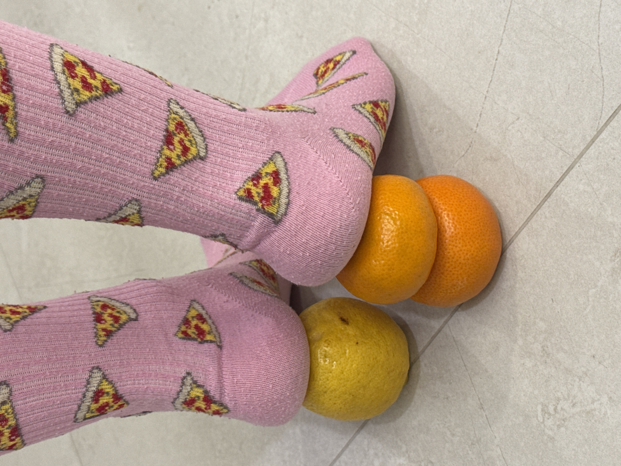 Two feet, clad in pink socks adorned with a repeating pattern of pizza slices, are positioned on a light gray tiled floor. One foot is pressed against a single yellow citrus fruit, resembling a lemon or lime. The other foot is pressing against two orange citrus fruits, likely tangerines or oranges. The fruits are round and appear fresh. The tiled floor has subtle variations in tone and visible grout lines, suggesting an indoor setting. The socks have a ribbed texture and show some minor pilling, indicating wear. The scene captures a casual, possibly playful interaction between the feet and the fruits. There is no visible text in the image. The location context provided is Chaikovsky, Russia.