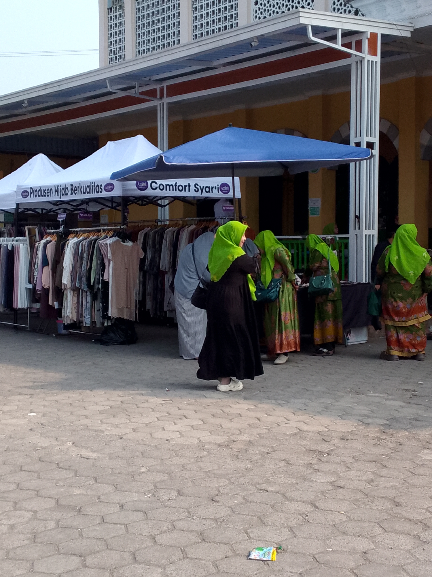 This image captures an outdoor market scene in Rajamandala Kulon, Indonesia, likely in front of a building with white pillars and an orange trim. A row of white and blue tents, branded with "Produsen Hijab Berkualitas" and "Comfort Syari", offers shade to vendors. Racks of women's clothing are displayed under the tents. Several women, dressed in hijabs and traditional attire, are browsing the merchandise. One woman in a black dress and green hijab is in the foreground, while others in green hijabs and colorful patterned dresses are in the background. The ground is paved with hexagonal grey stones. A small, colorful item, possibly a snack wrapper or pamphlet, lies on the ground in the lower right corner. The overall atmosphere suggests a bustling marketplace focused on modest fashion.